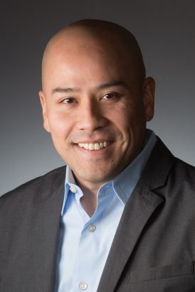 sam has a shaved head and medium complexion and wears a blue collared shirt and grey sport coat