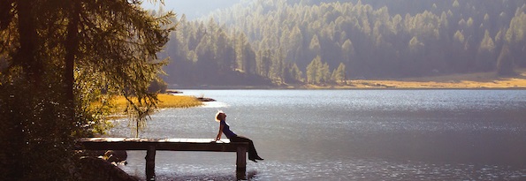 sillouette of a person with their legs hanging off a dock in a lake with pine trees behind them