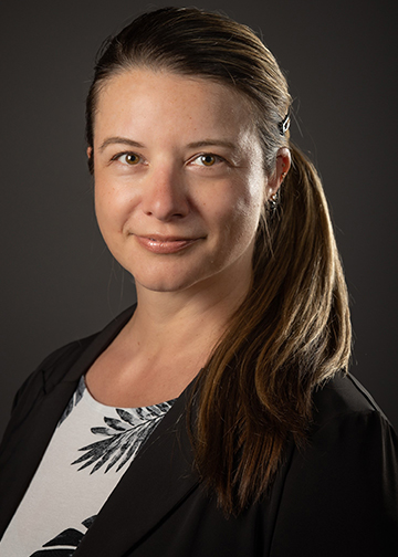 rachel has a long brown ponytail and wears a black jacket over a black and white palmtree print shirt