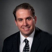 Michael Cassara has short brown hair and is wearing a black suit, with a white shirt, and black and white tie.