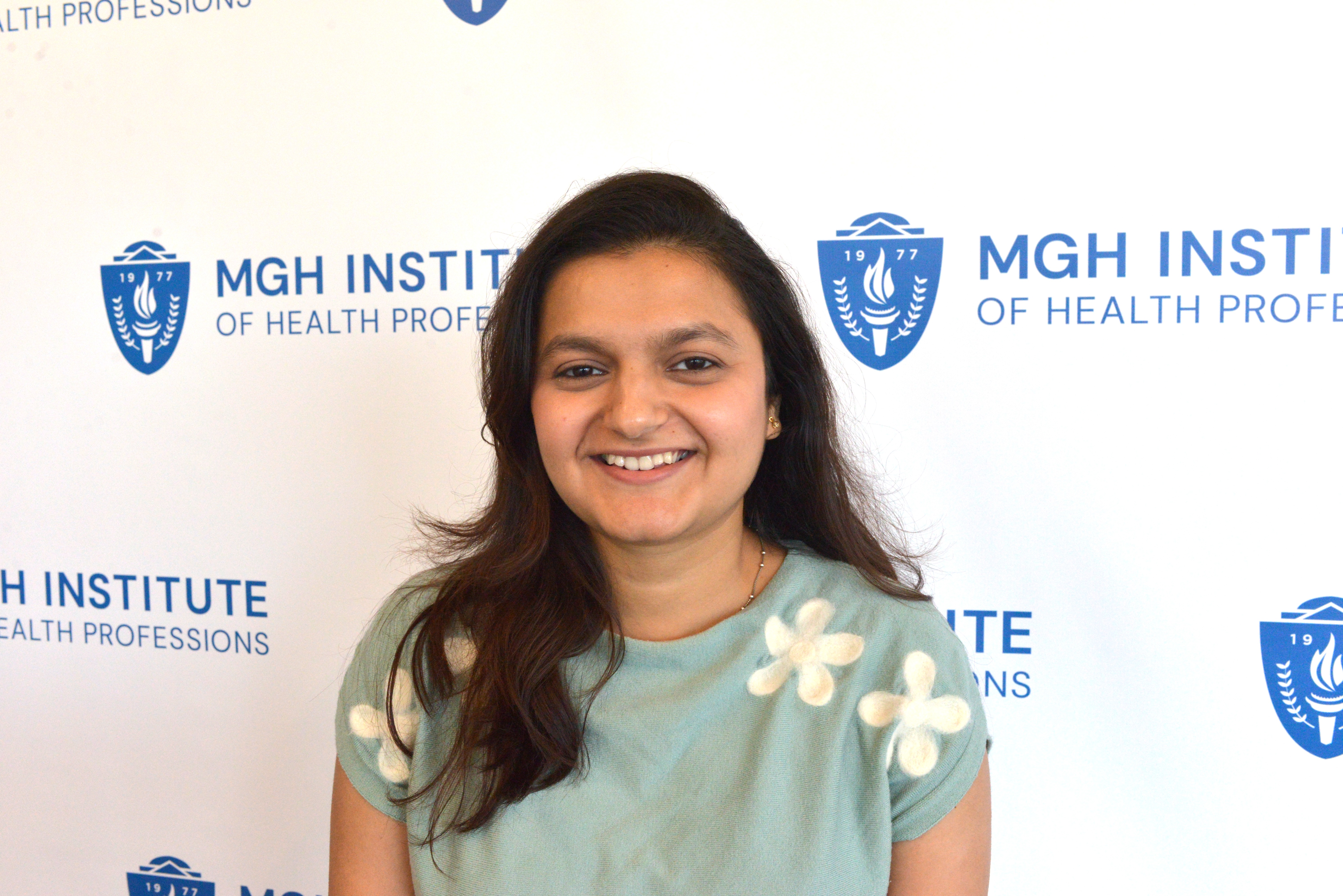Woman with long black hair wearing a light blue shirt with a flower design on it standing in front of a background featuring the MGHIHP logo