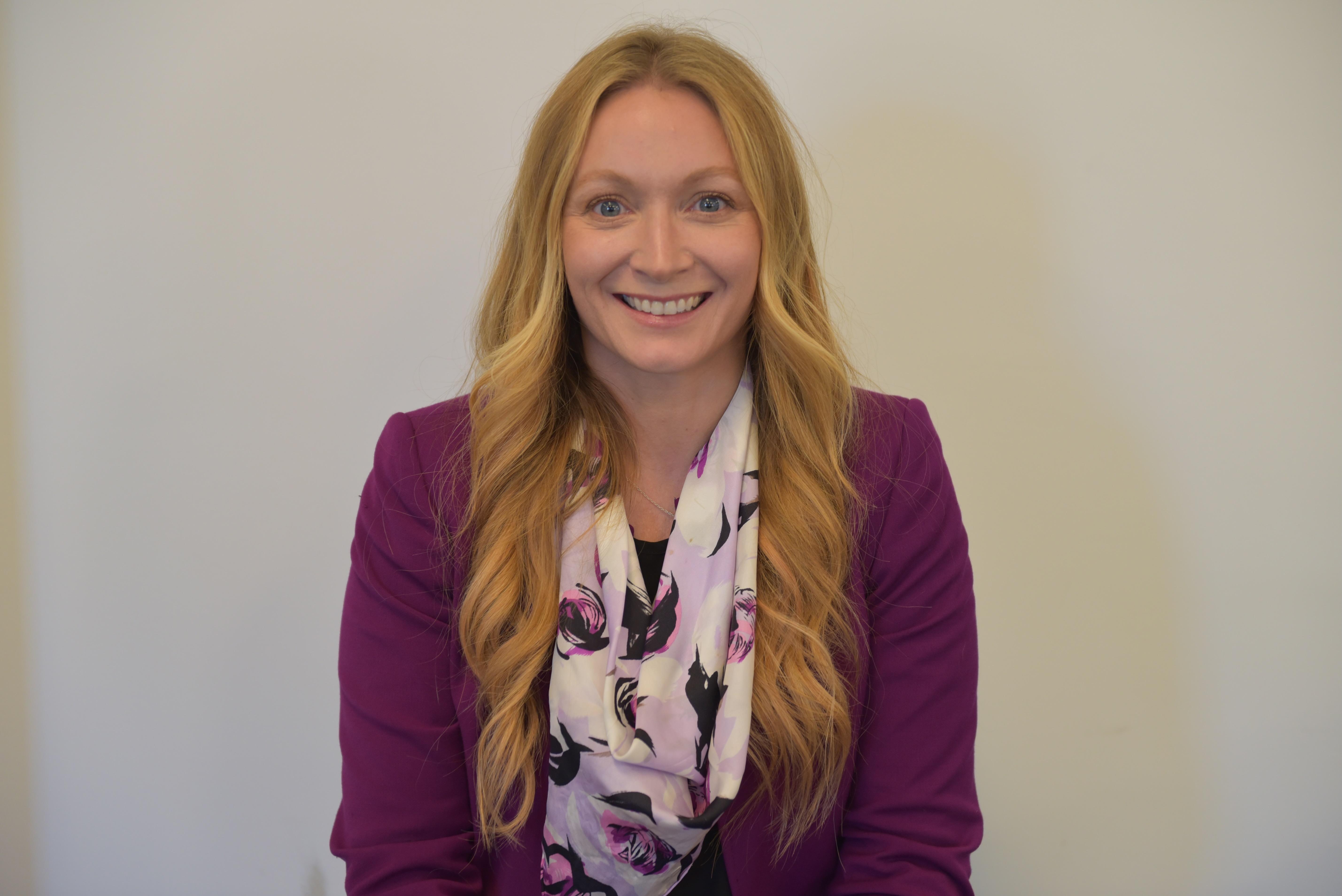Headshot of woman with long blonde hair standing in front of solid white background wearing a light purple scarf and dark purple jacket