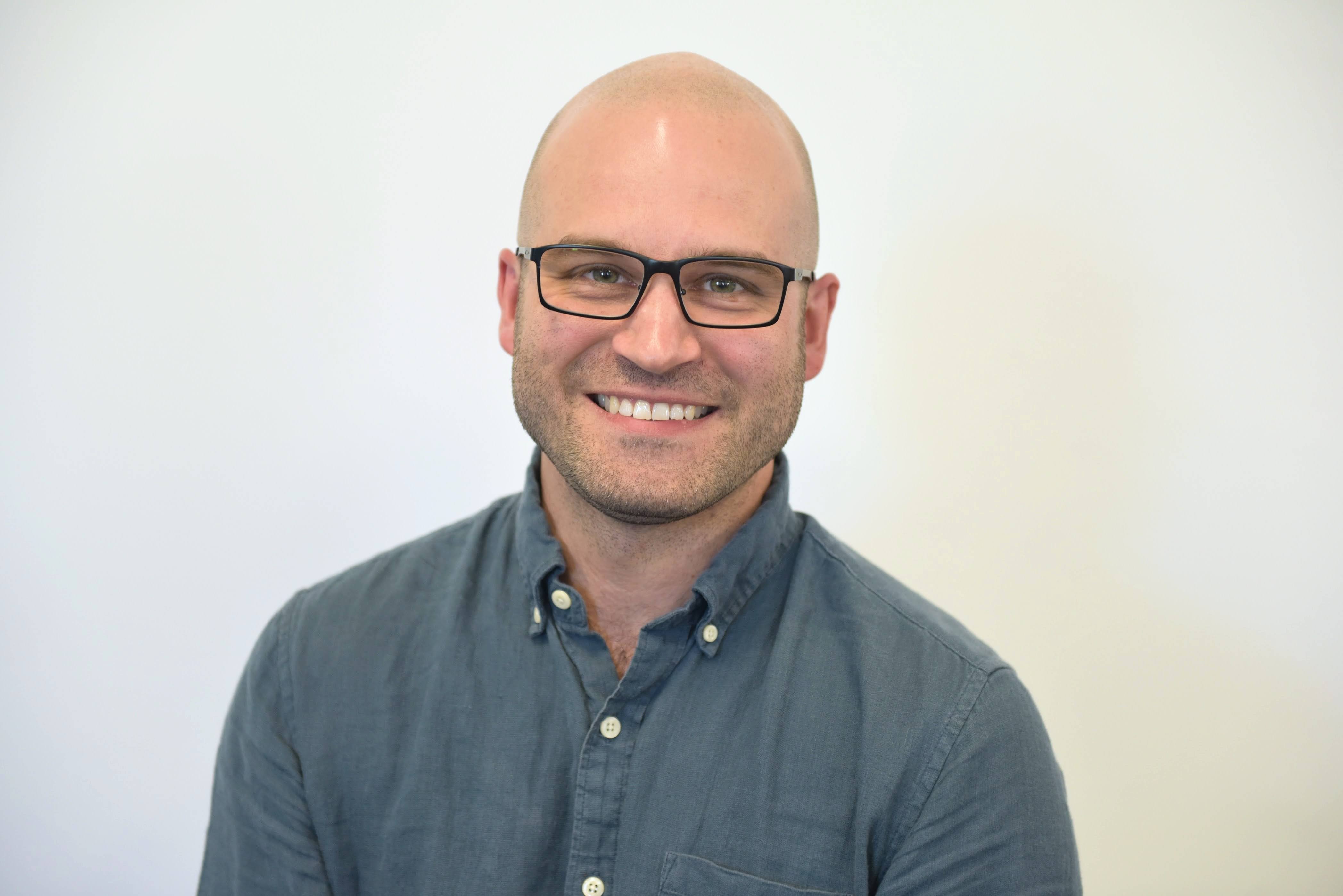 Headshot of man against a solid white background blue button-down shirt and glasses.