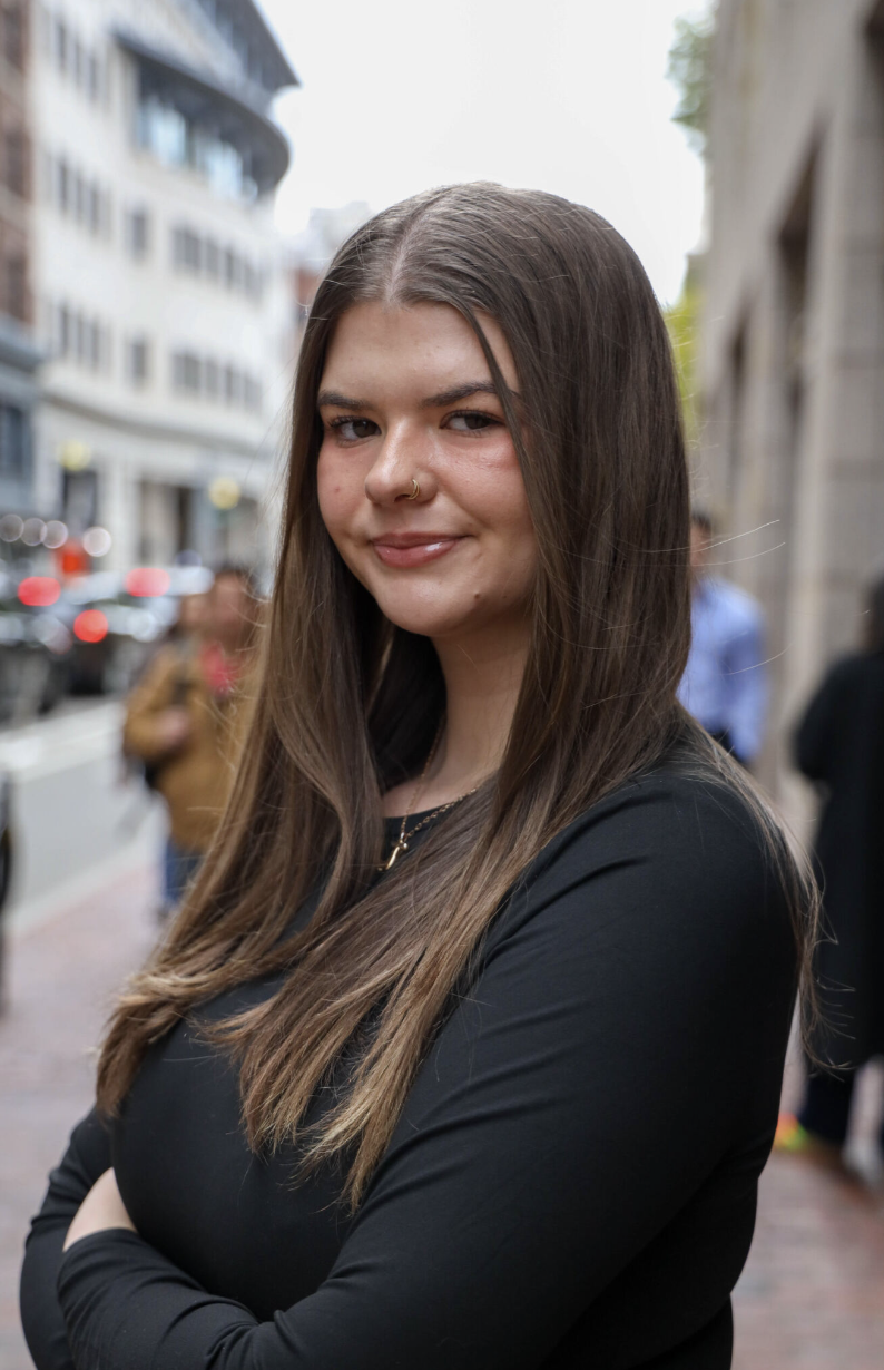 Headshot of woman with long brown hair wearing a black shirt in front of a city street in the background