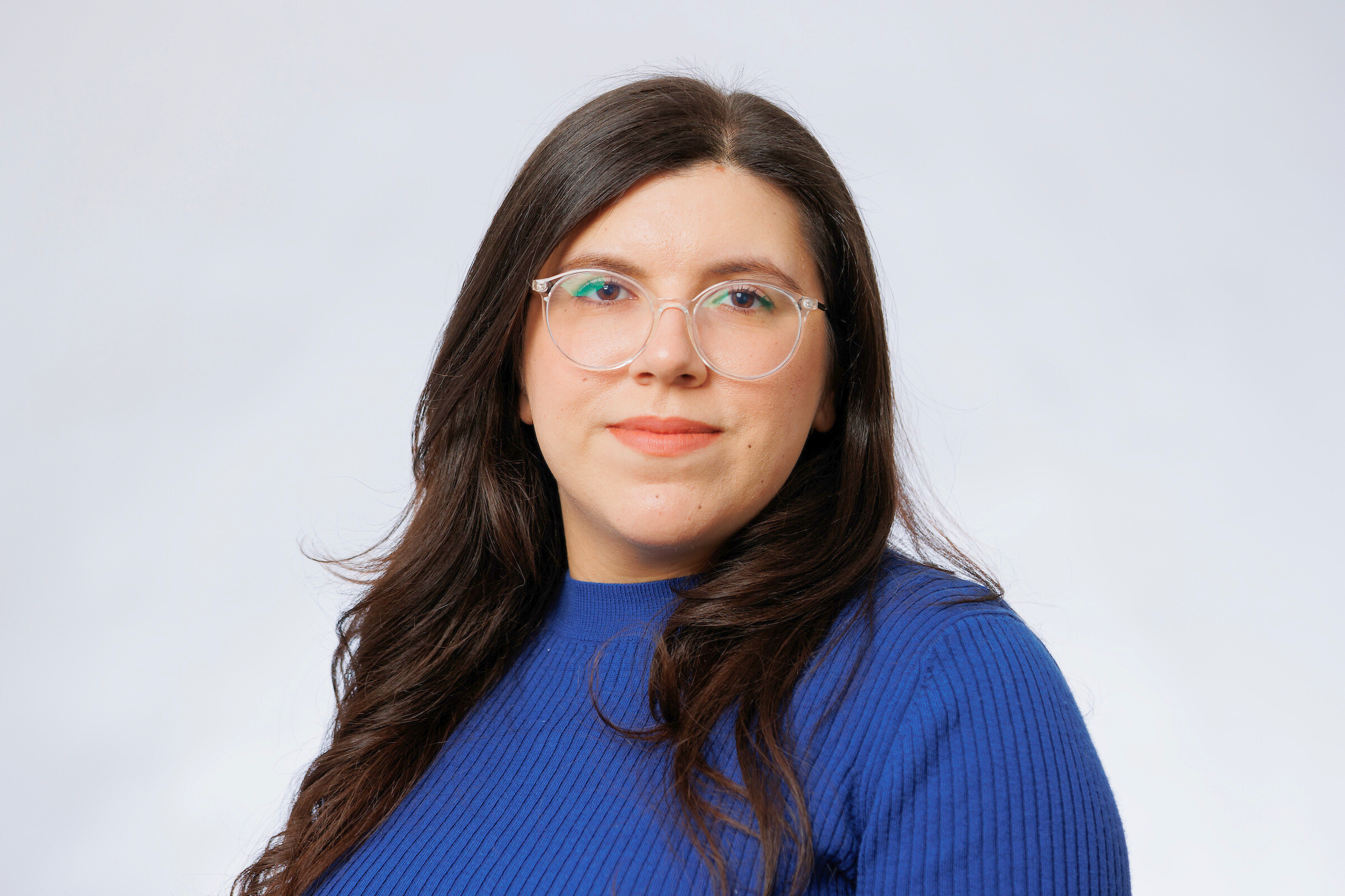 Headshot of woman with long black hair wearing glasses and a blue shirt against a solid gray background