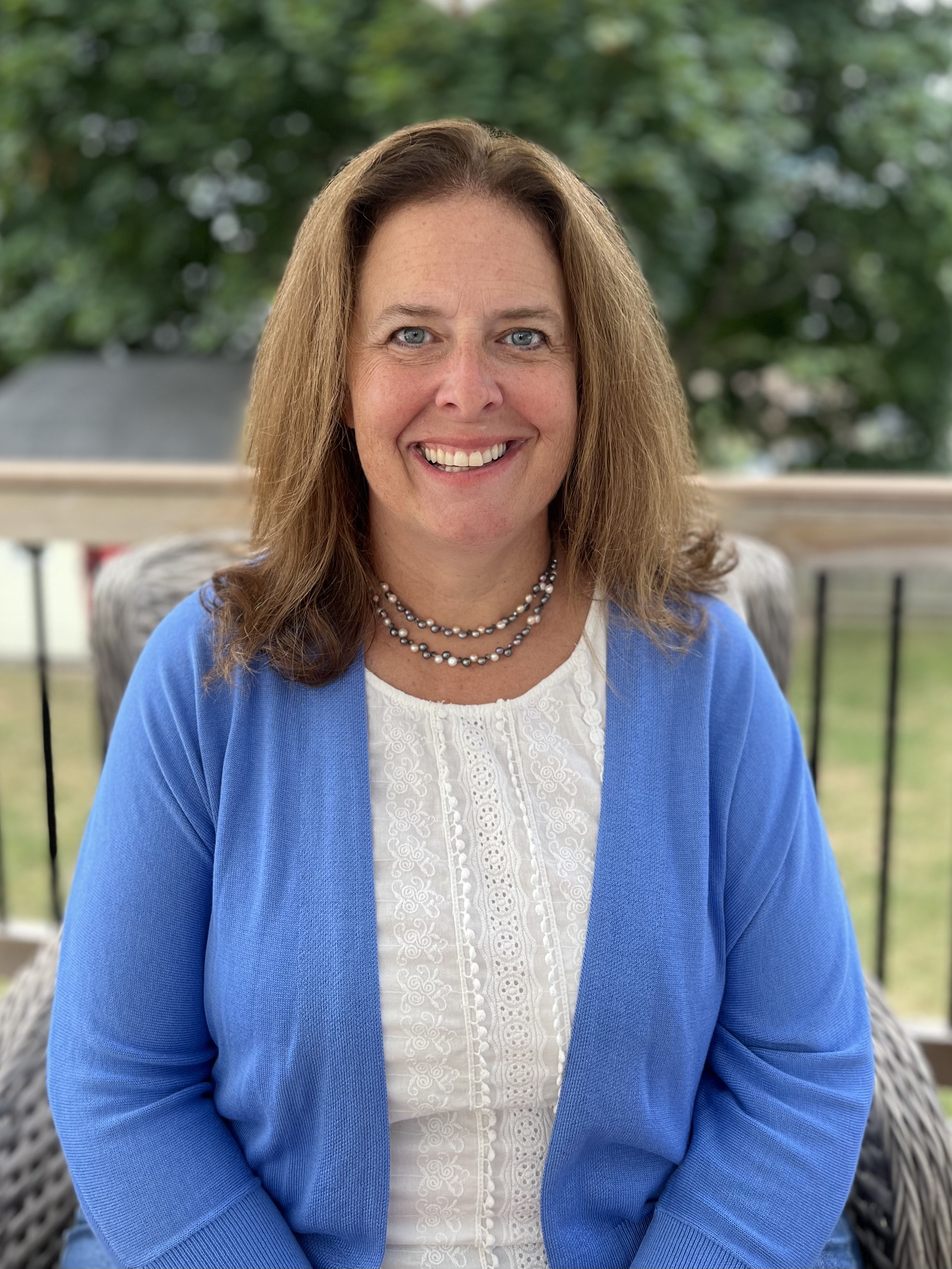 Headshot of woman with long brown hair wearing a white shirt, a blue sweater, and a necklace sitting on a balcony overlooking a lawn.
