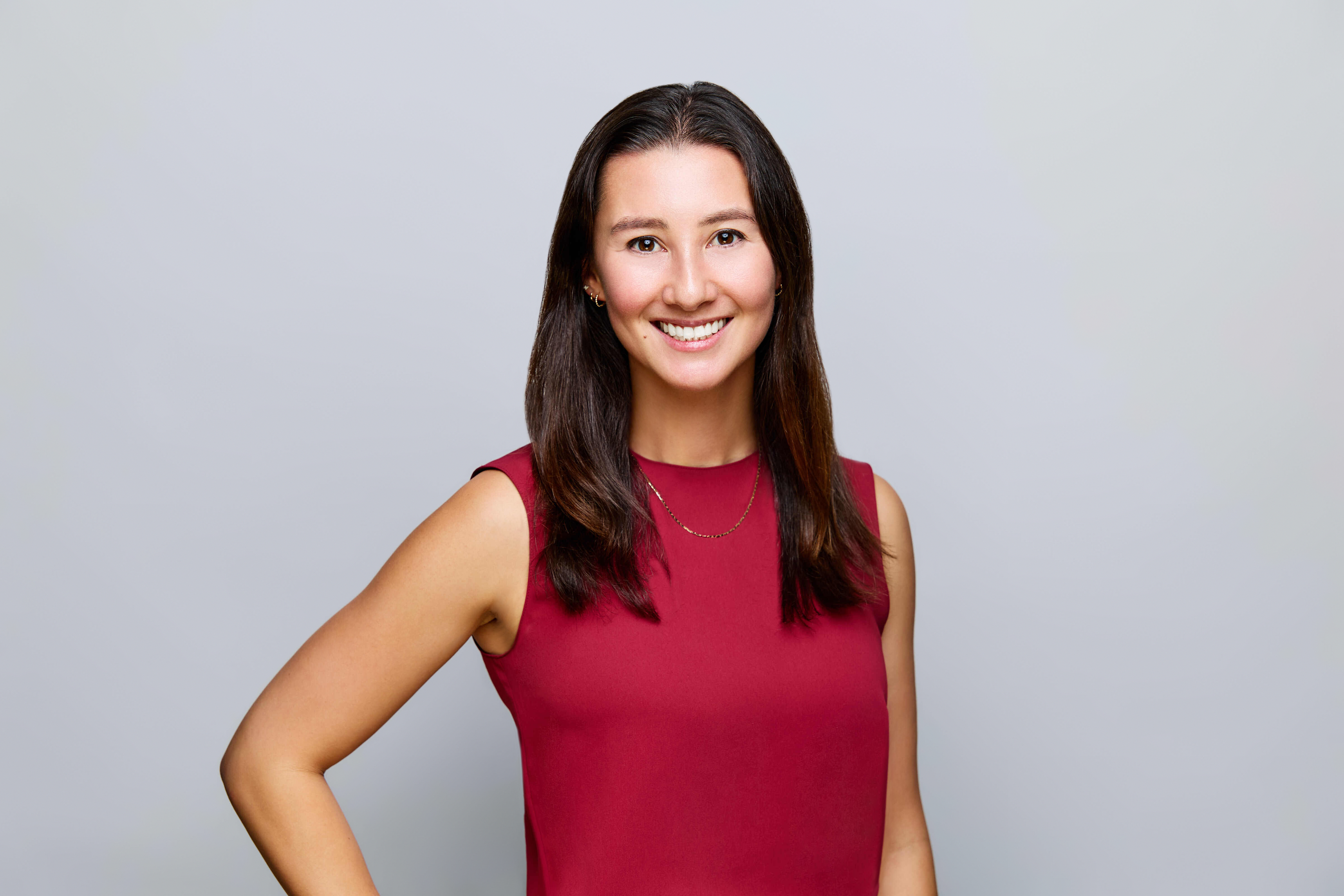Headshot of woman with long dark hair wearing a red shirt against a gray background.
