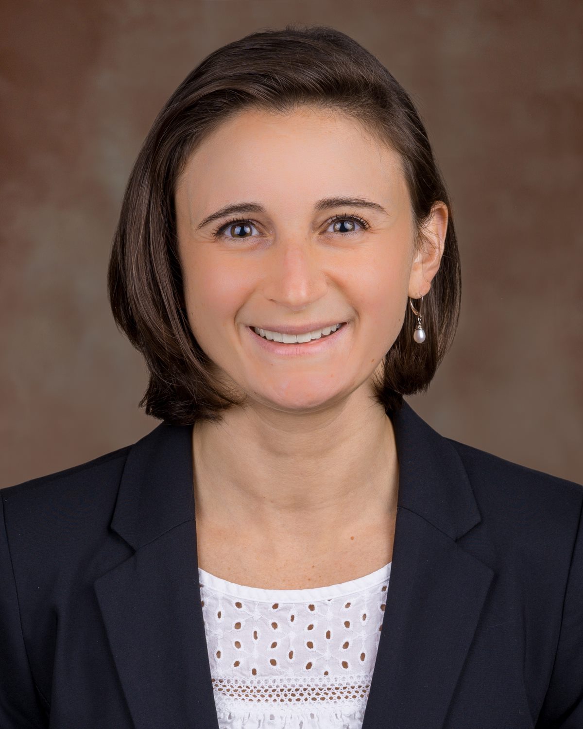 Headshot of woman wearing white shirt and black jacket against a brown backgroud