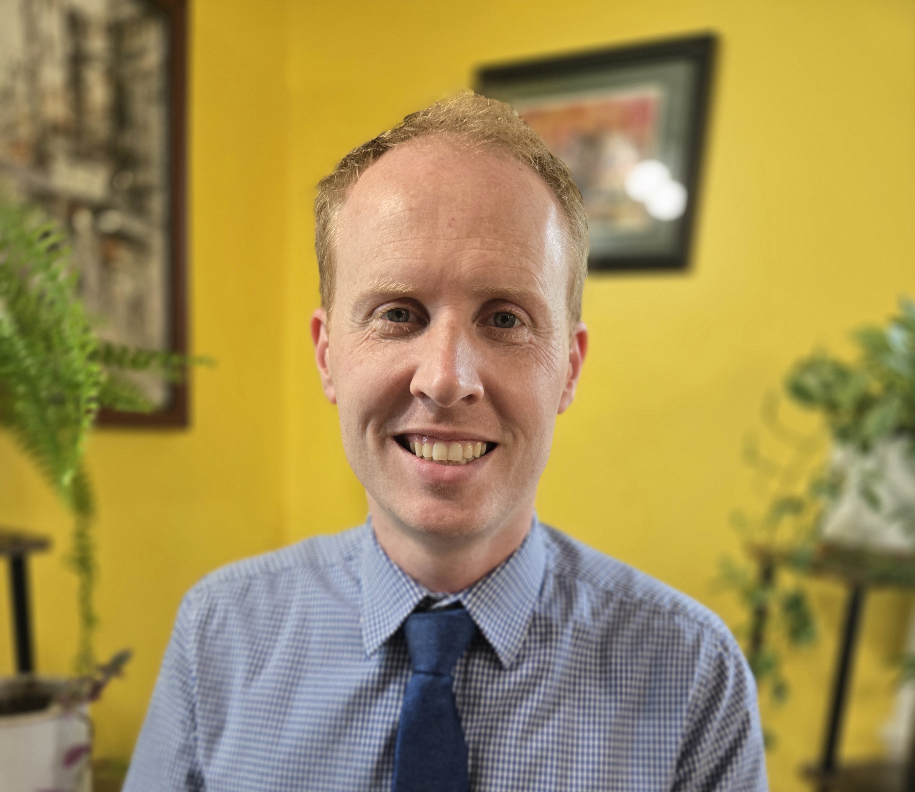 Headshot of Christopher Bodden wearing a blue button up shirt and blue tye in front of a yellow wall with paintings hung in the background.