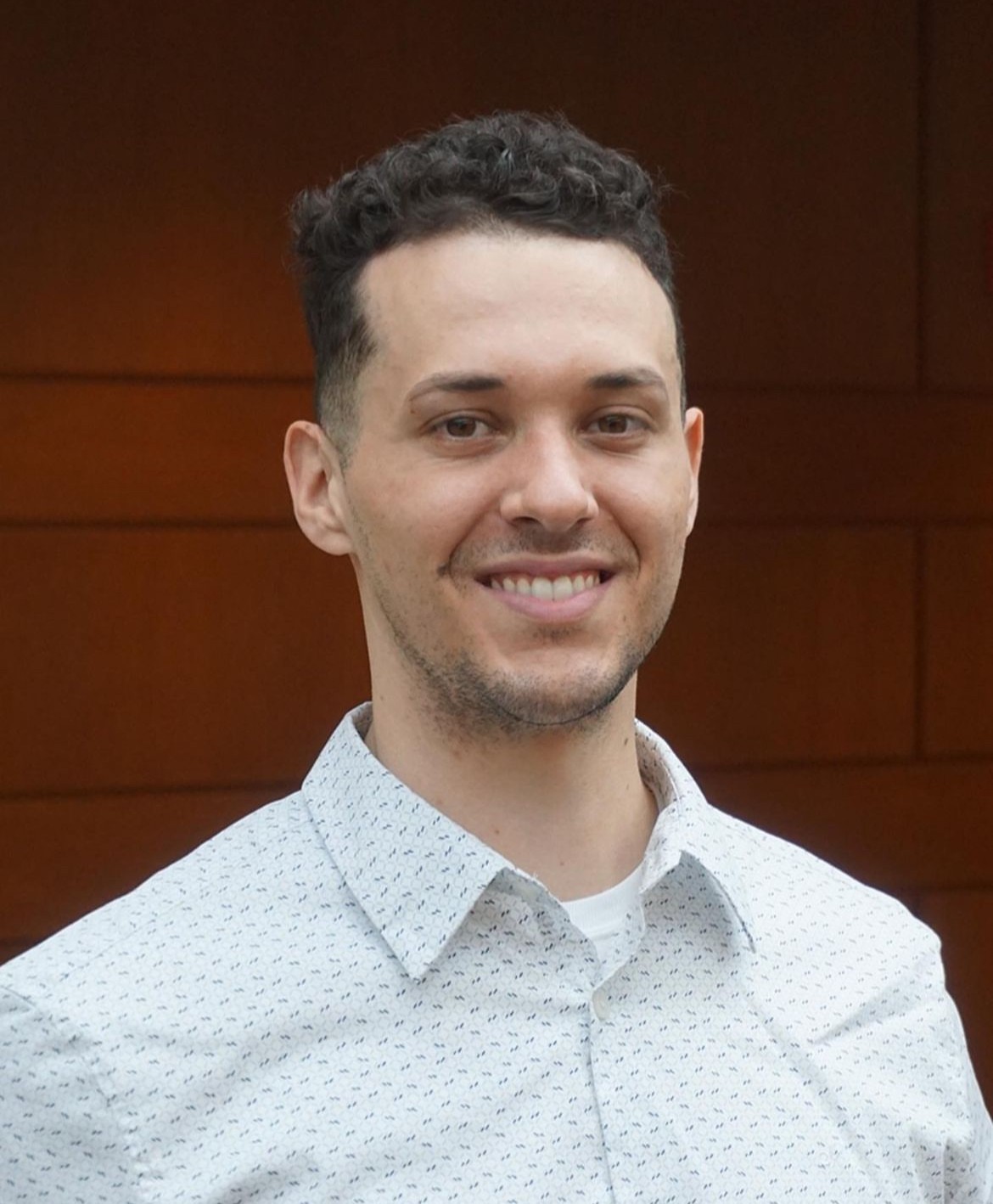 Man with short dark hair wearing a white patterned collared shirt in front of a red background