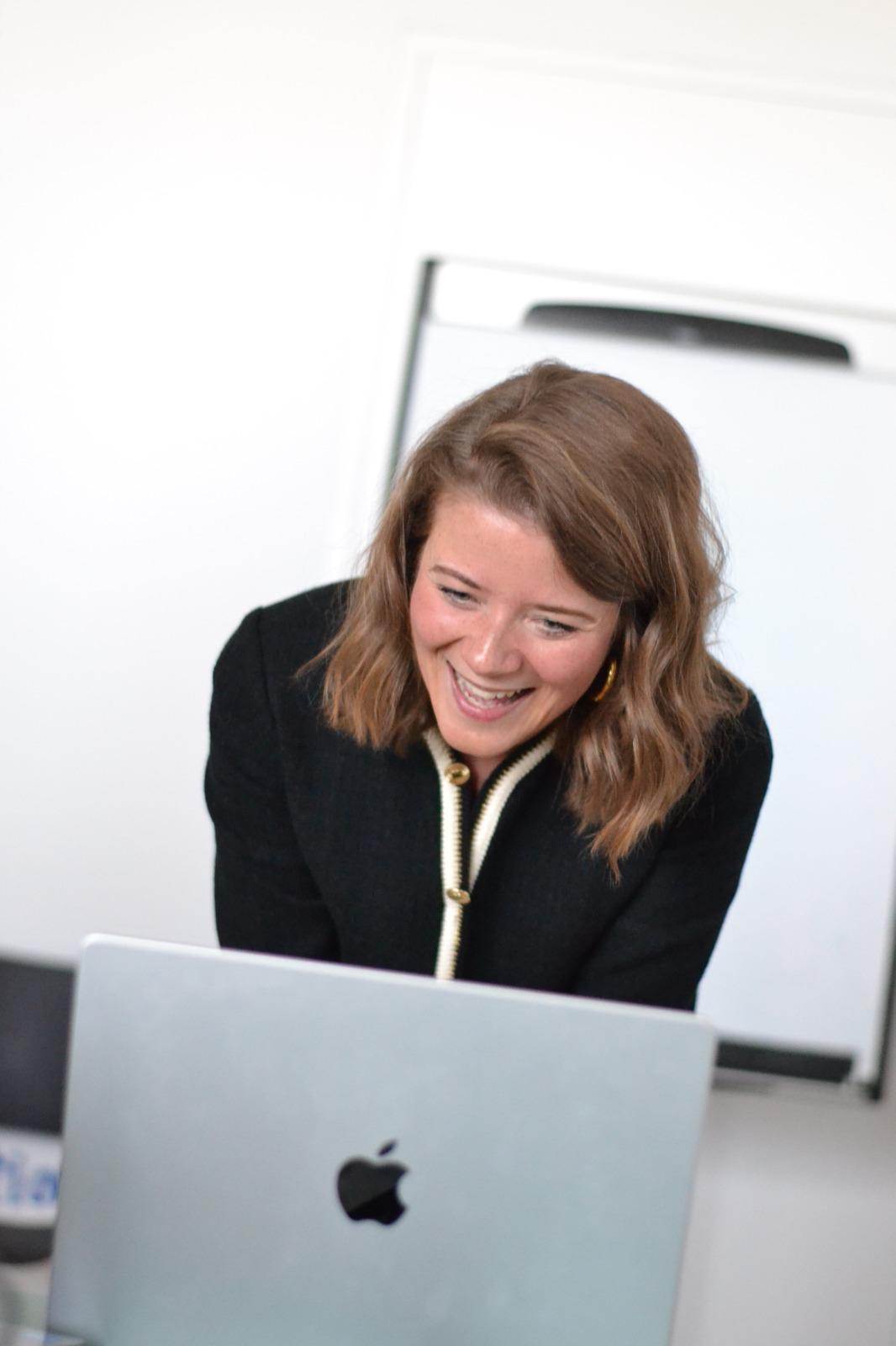 Photo of Weyland with shoulder length brown hair standing in front of a Mac laptop wearing a black top