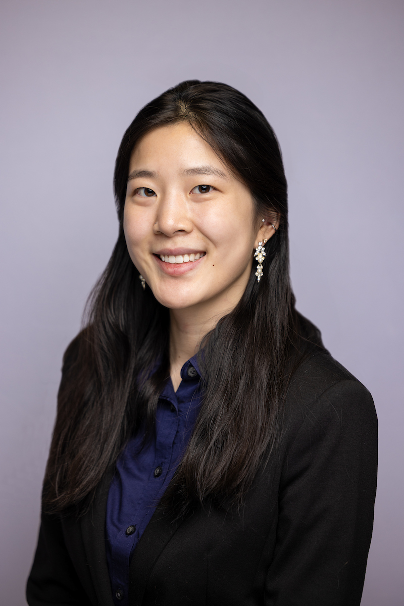 Headshot of woman with long black hair wearing a blue button-down shirt, a black jacket, and earings