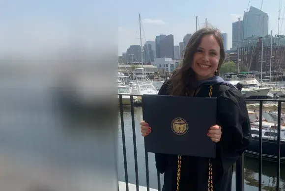 A woman wearing a graduation gown holds a diploma in front of a background of sailboats