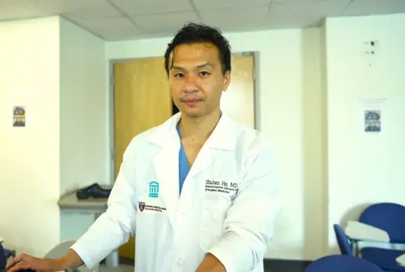 Doctor in lab coat looking into camera; coat has name and hosital affiliation 