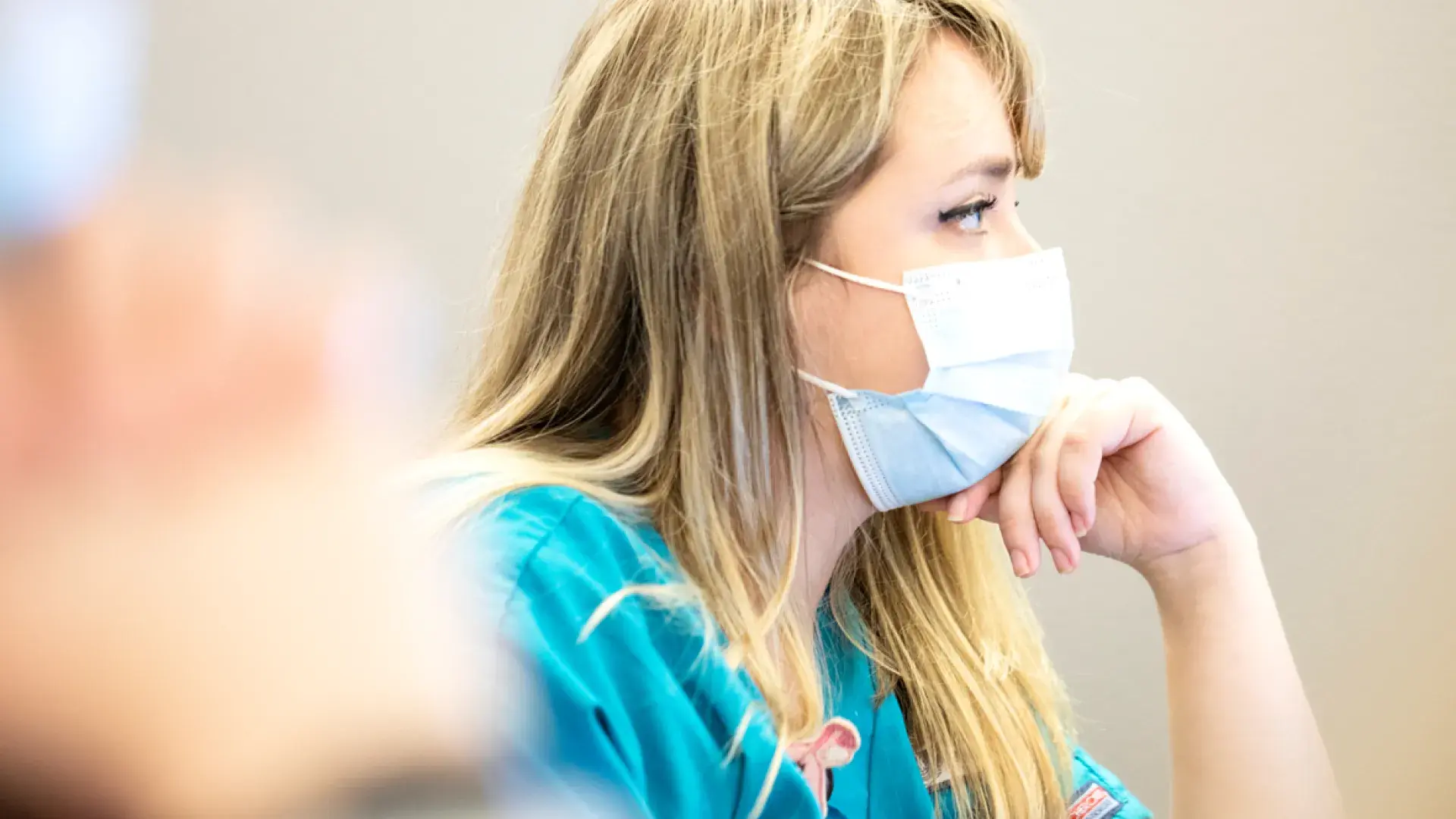 A woman sitting with a mask on and her chin resting on her hand