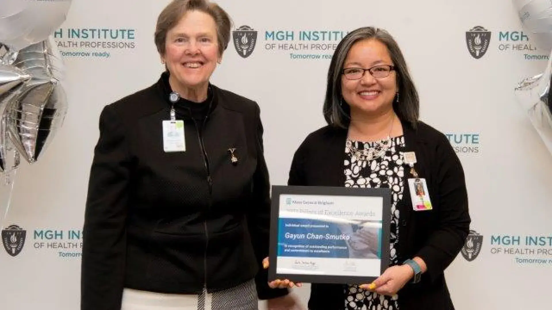 Gayun holds award in front of banner and stands with paula