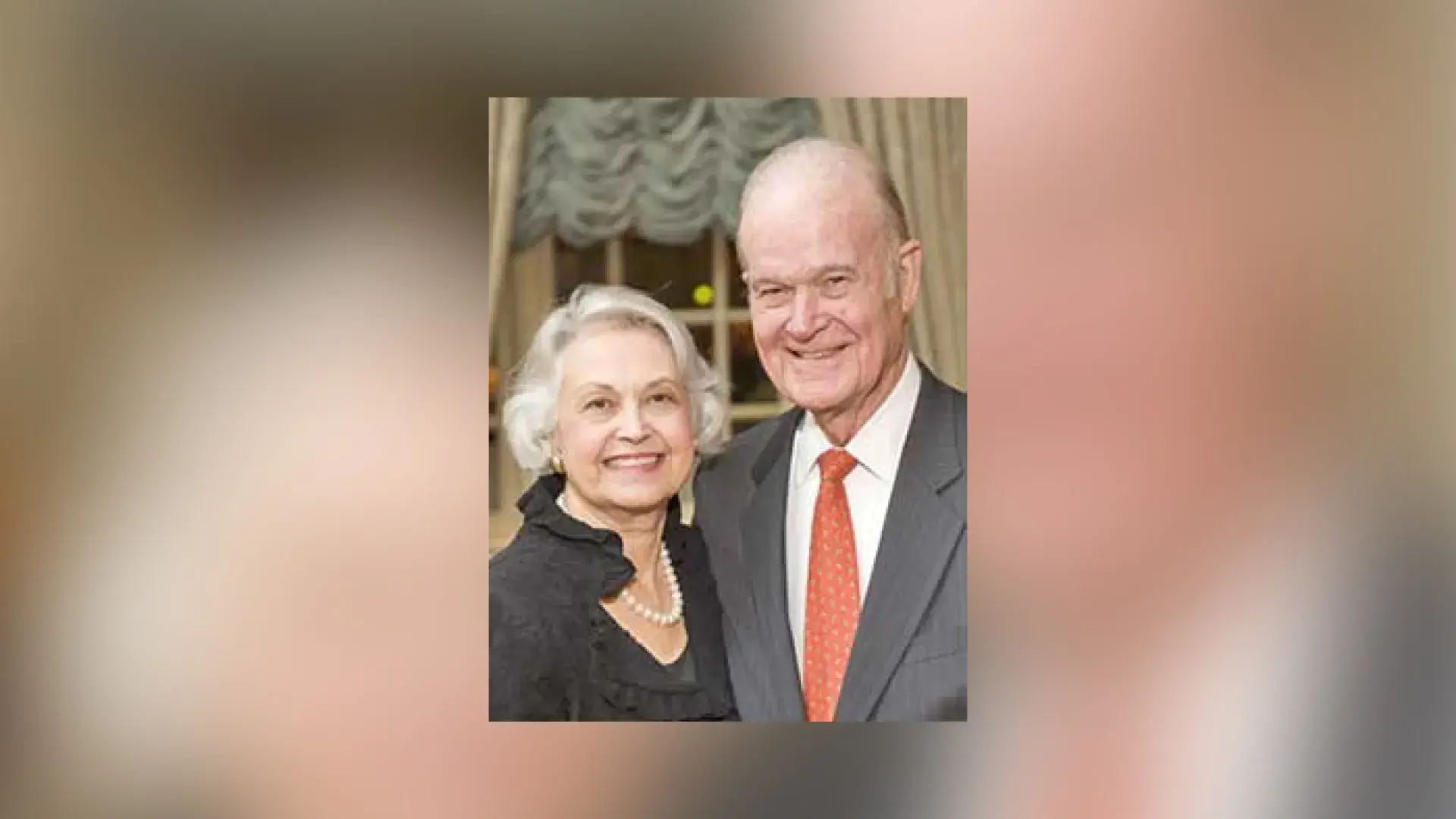 charles wears a grey suit and red tie, ann wears a black shirt with pearls