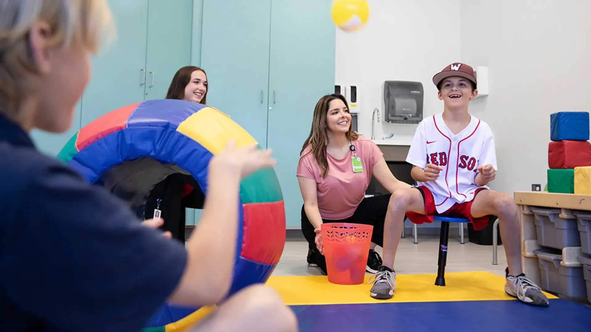 child in red sox jersey balancing on a stool smiles as a person in the foreground tosses a yellow ball to him