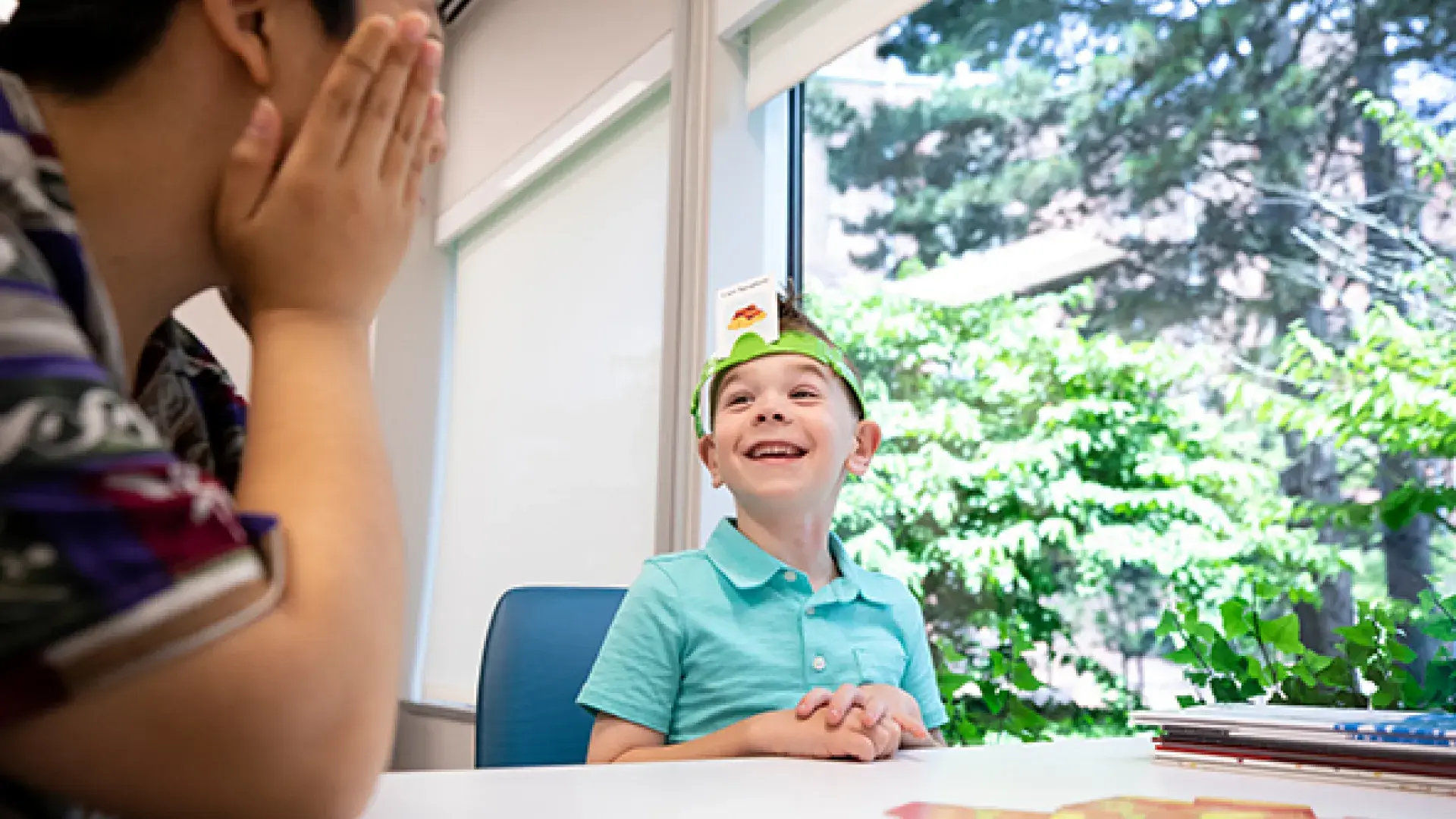 6 year old child smiles, sits at table playing a guessing game - a card on headband around his head says "i am spaghetti"