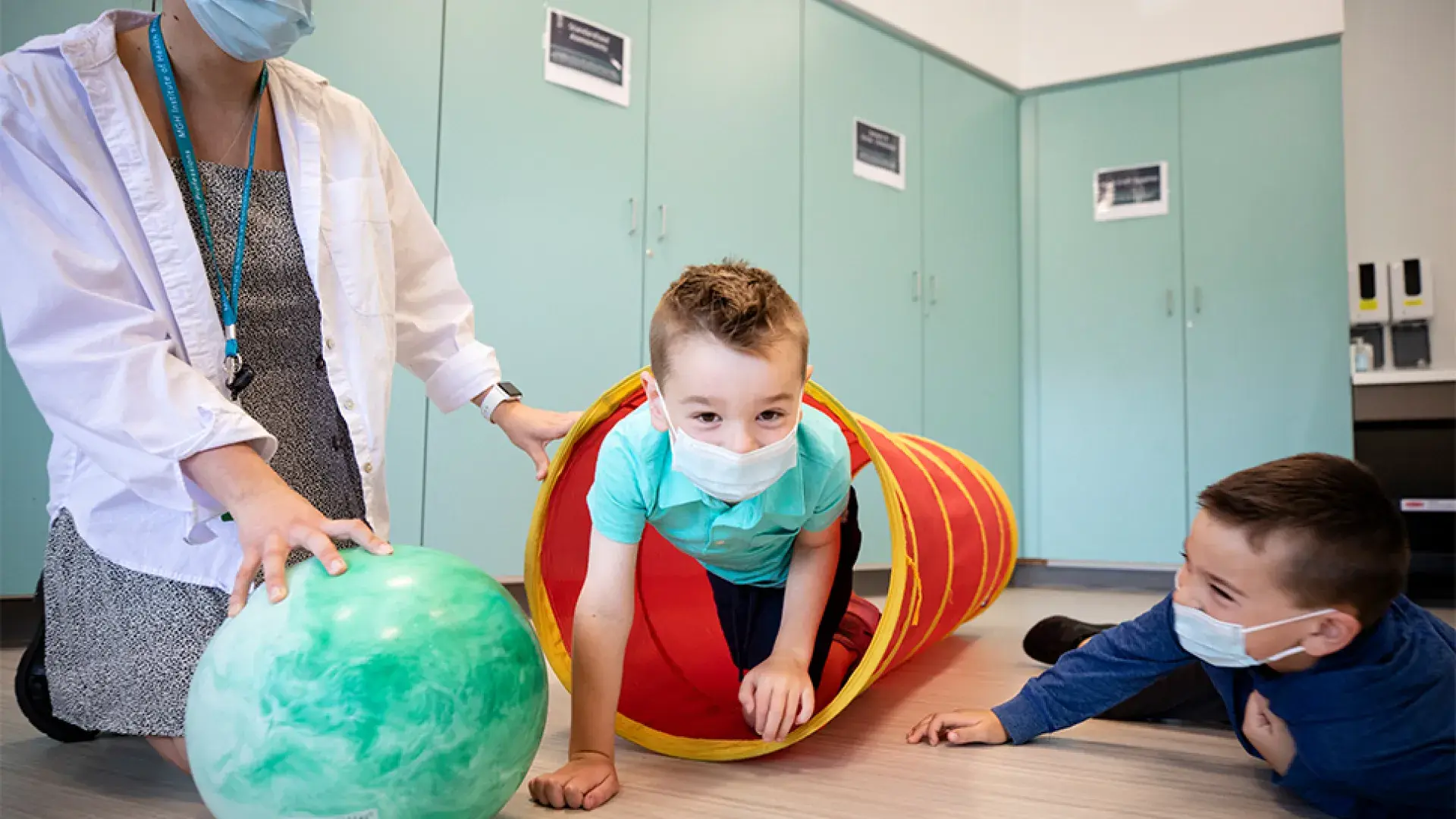 a little boy in a mask is helped by an IHP student to crawl through a cloth tube while his brother looks on