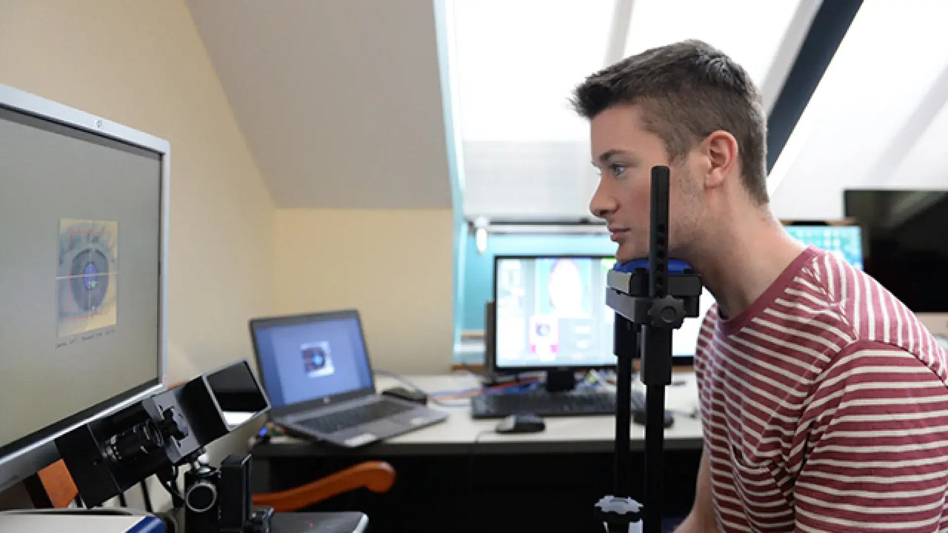 A man sits in front of a computer screen with his head on a stand device while his eye is scanned and displayed on the monitor.