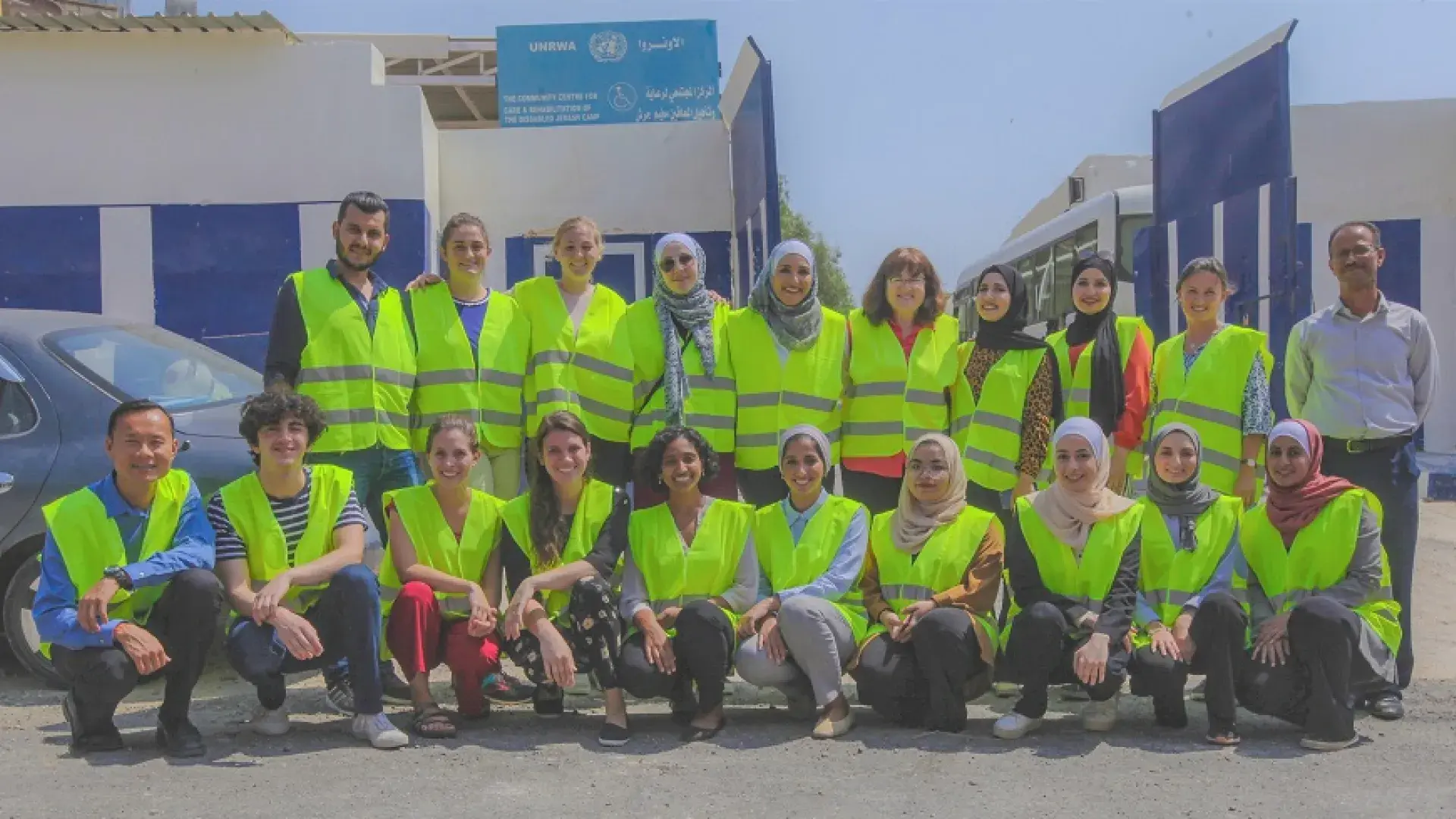 20 people pose in front of a building with arabic writing on it, they wear yellow vests