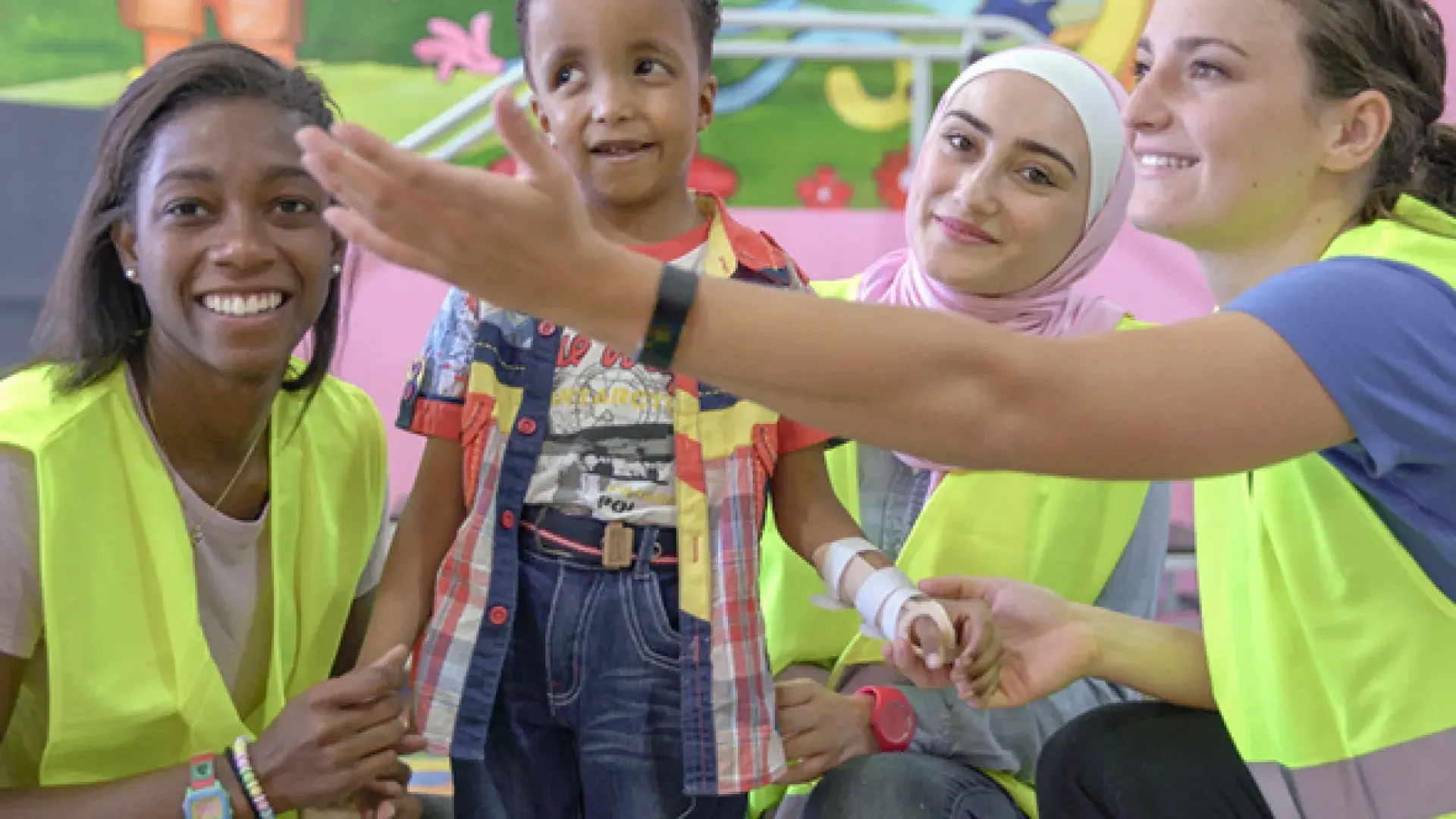 woman gestures to a little boy in a cast while two other women in yellow vests hold his hands