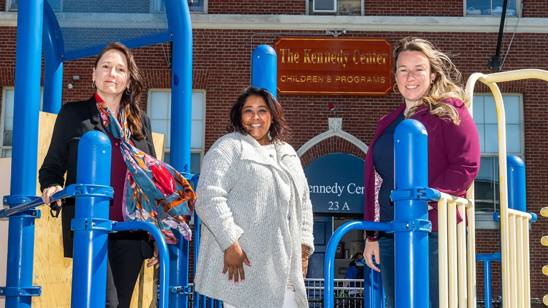 three women stand on a jungle gym with kennedy center sign in the background