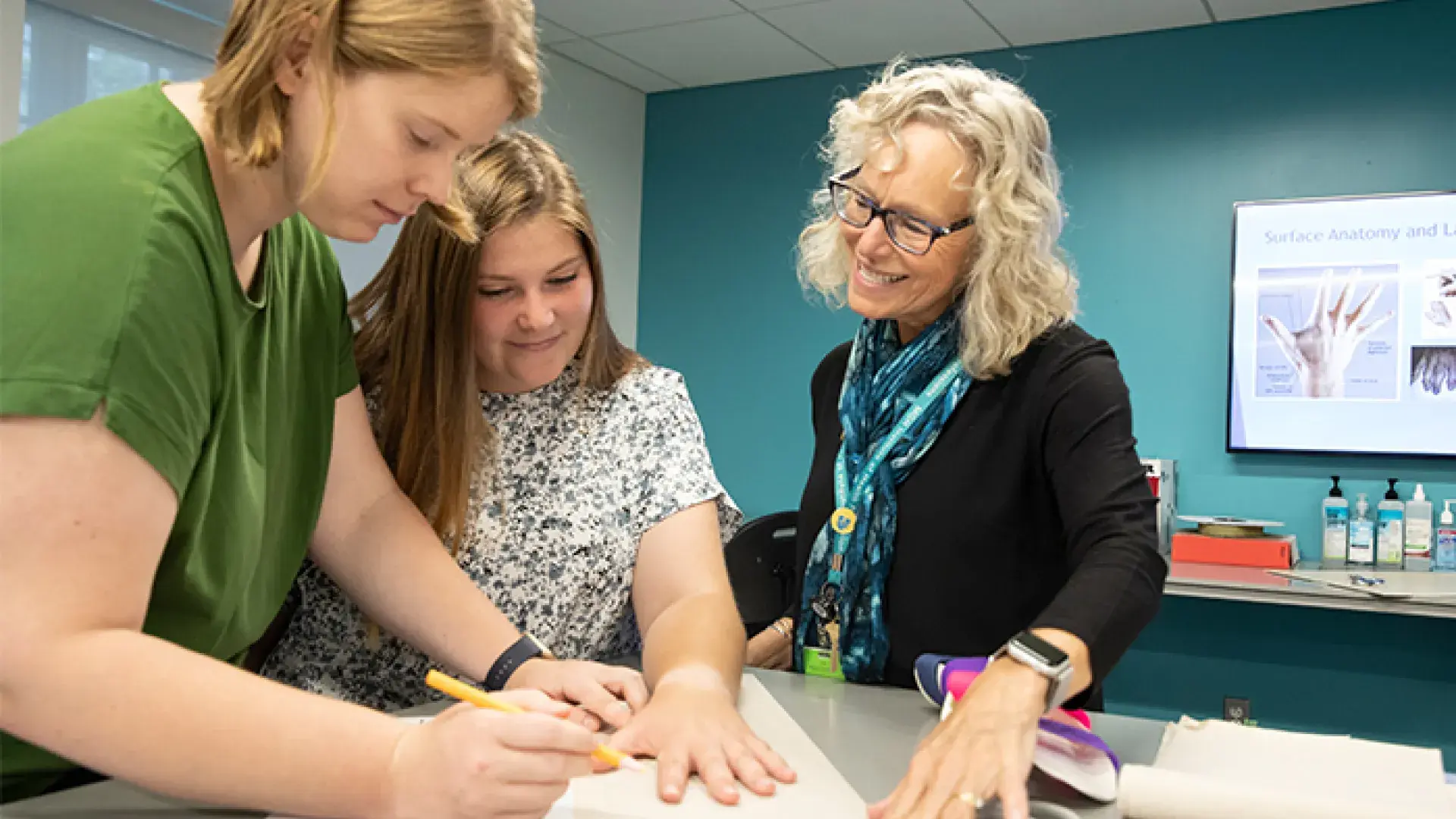 young woman with short strawberry blond hair traces another woman's hand on paper while another woman looks on