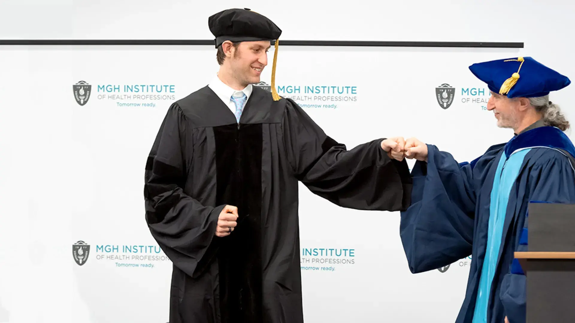 Man in graduation robe and cap fist bumps another man in blue graduation attire