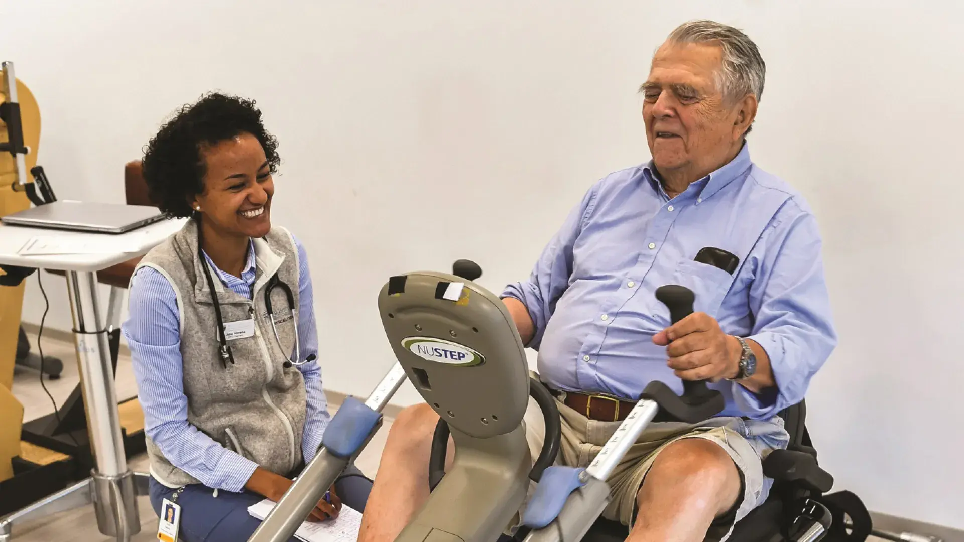 young woman with a stethoscope sits by elderly man on recumbent bike