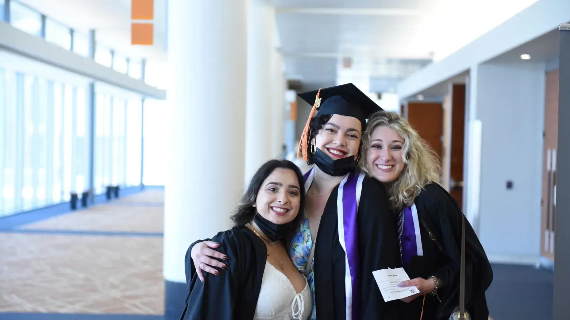 Three women in black graduation robes stand with their arms around each other smiling. One wears a graduation cap.