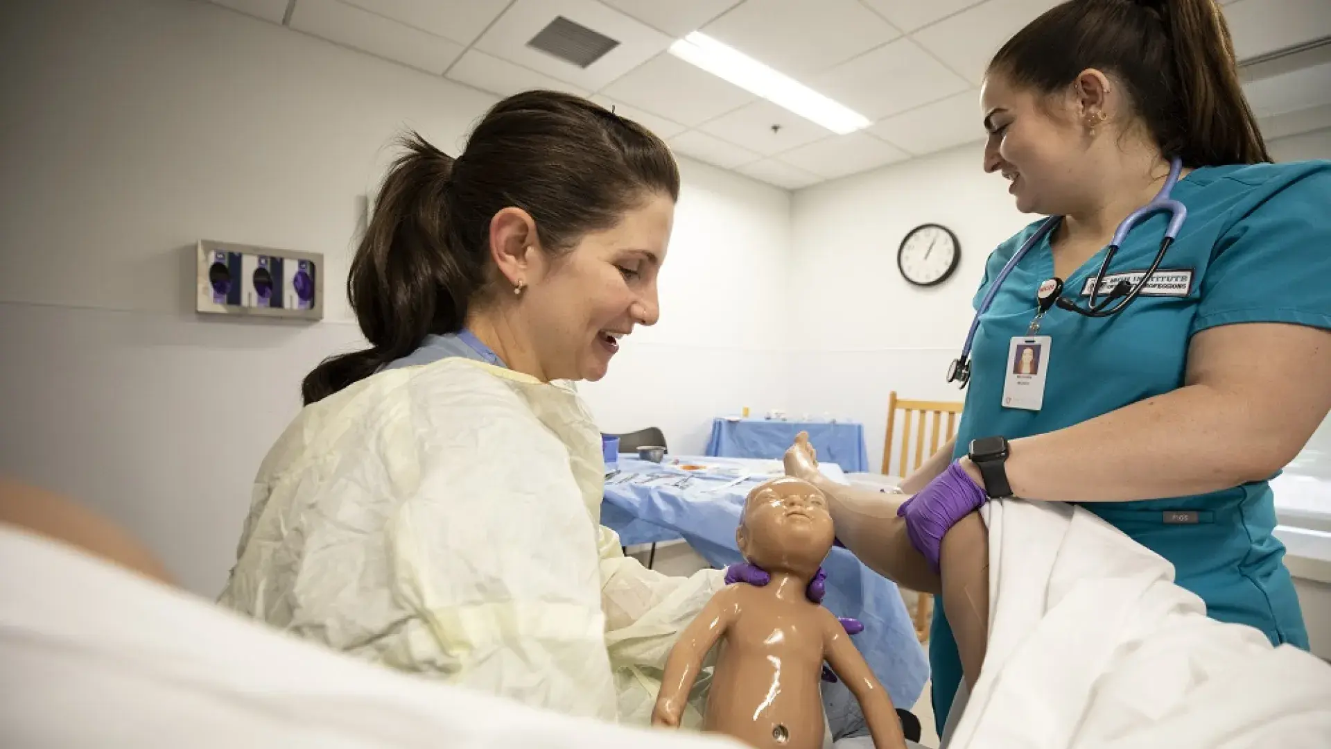 woman in gown holds baby manikin covered in liquid while another holds the manikin mom's leg