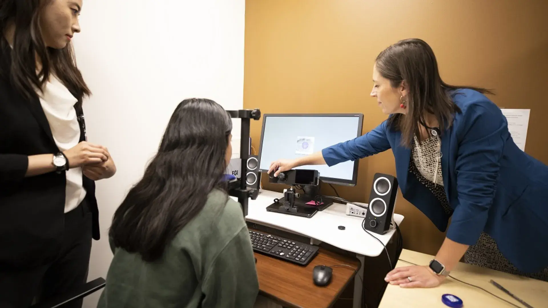 woman rests chin on black device and looks at a screen and camera while a woman points at the screen