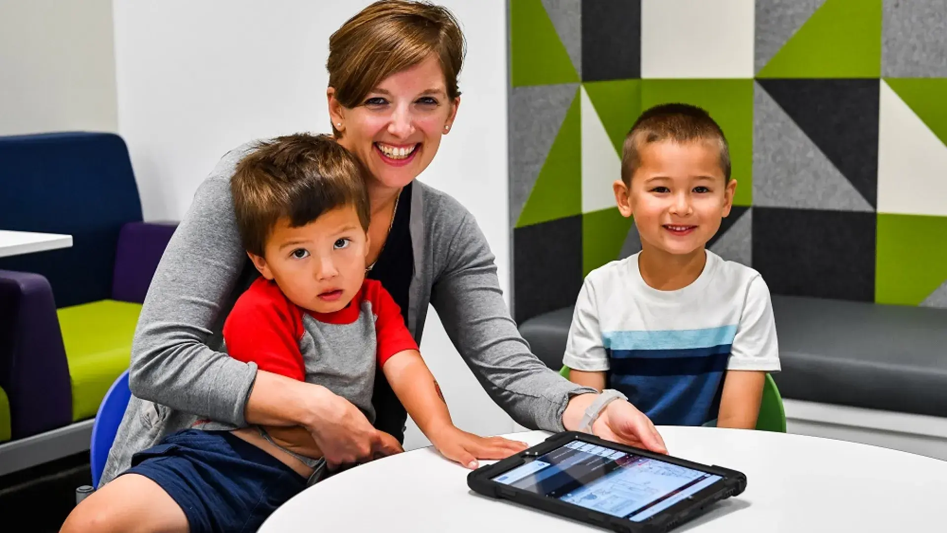 two young boys smile at the camera one sits on a woman's lap who holds a tablet