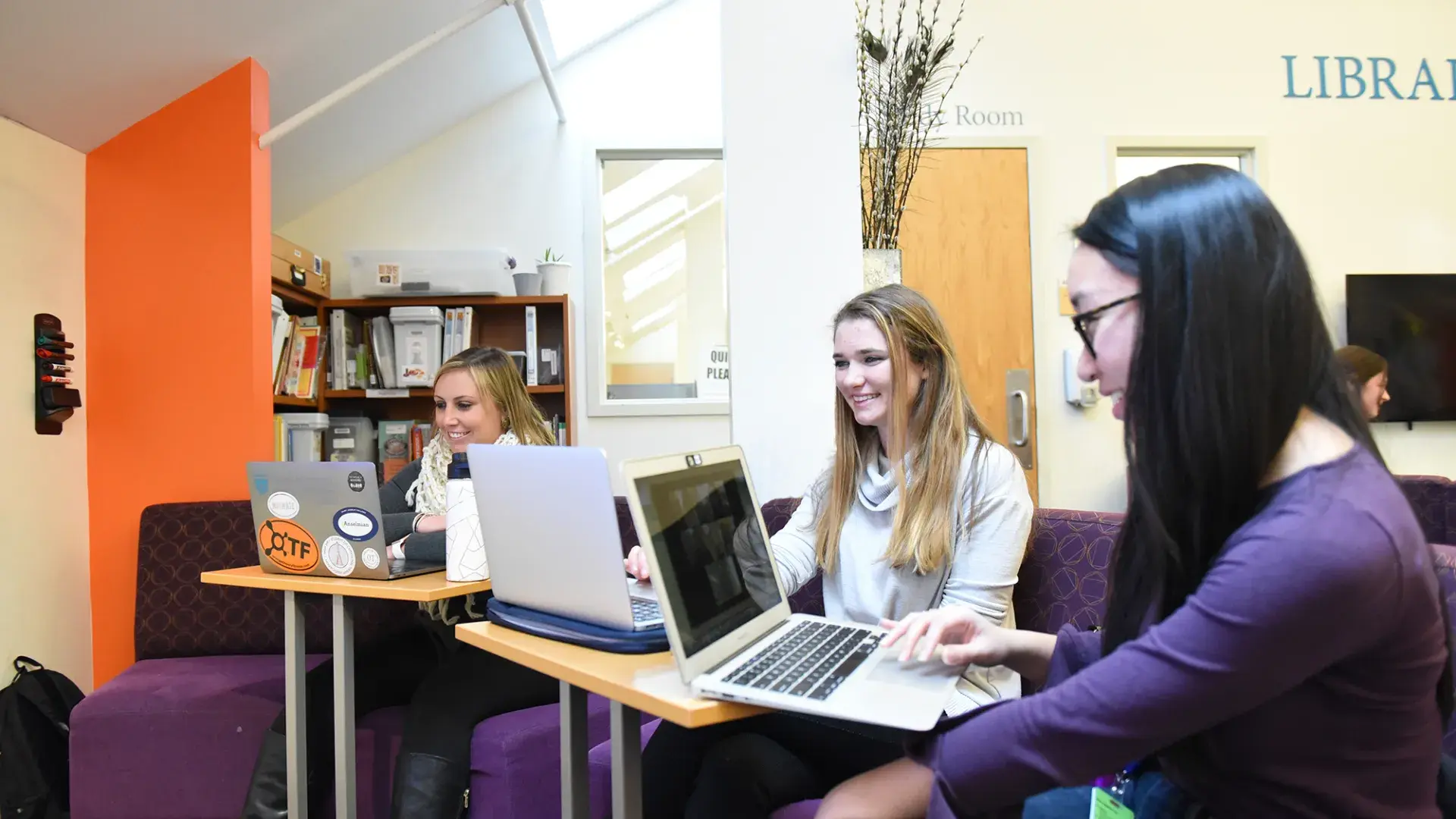 three women sit on a couch with laptops on tables