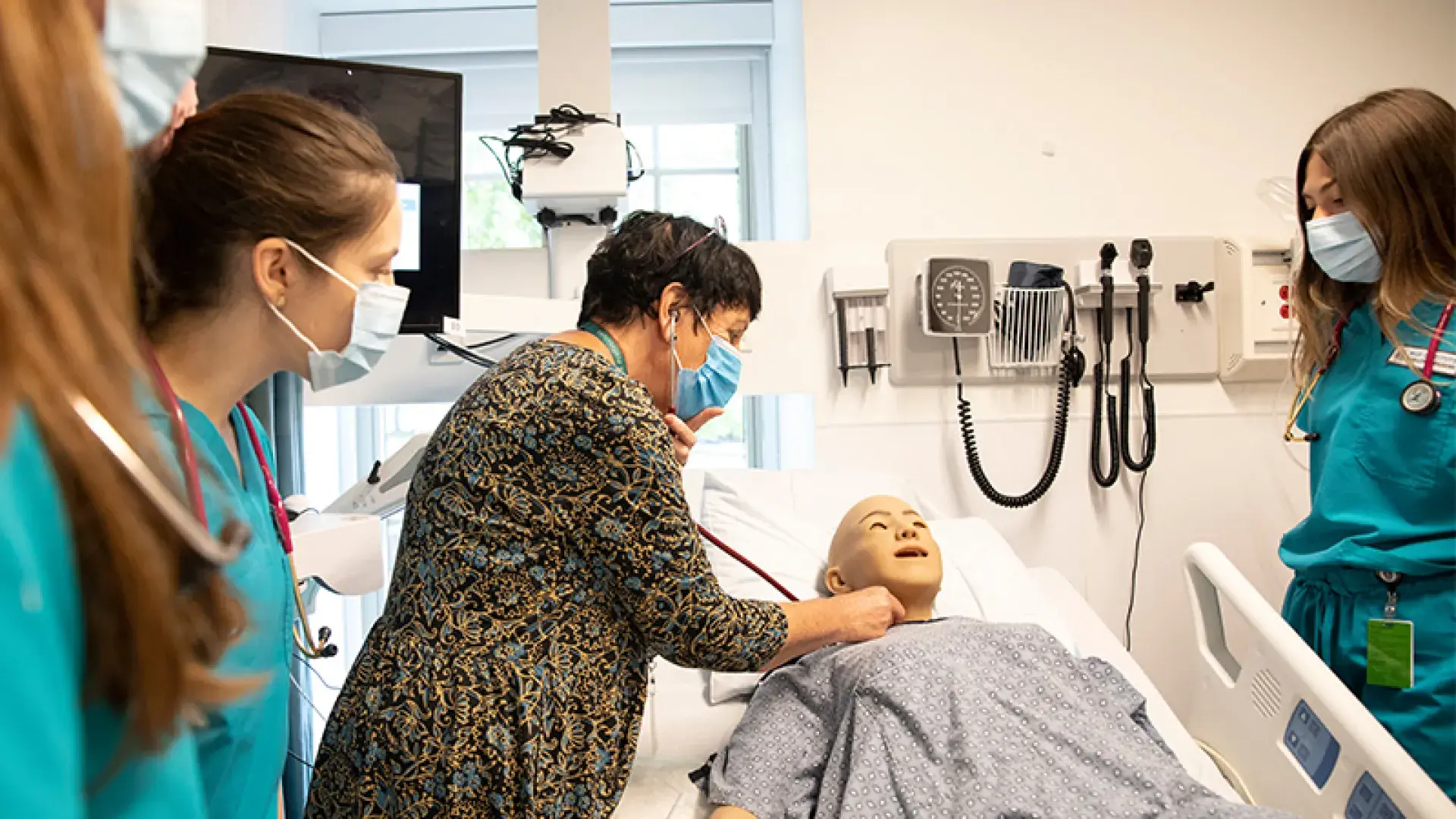 faculty member listens to the neck of a manikin as students in scrubs look on