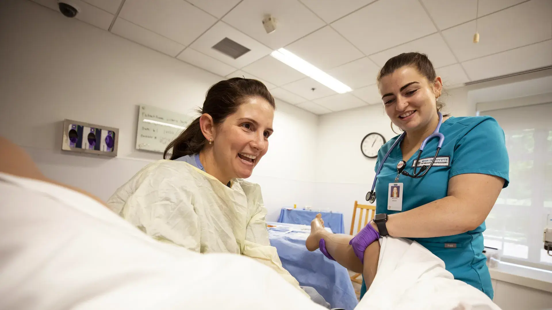 woman in scrubs holds a manikin leg while another women in a gown delivers a manikin baby