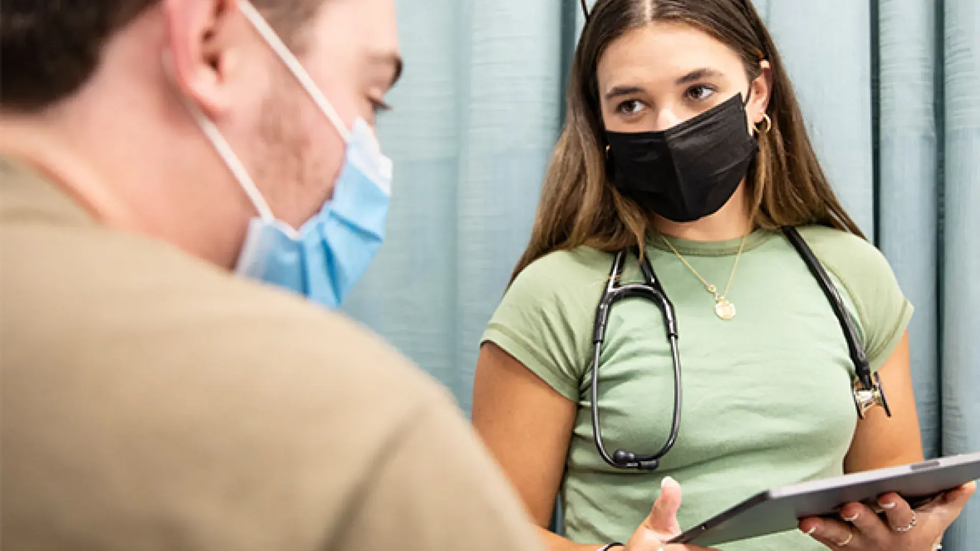 one student with a stethoscope takes a history  from another student using an ipad