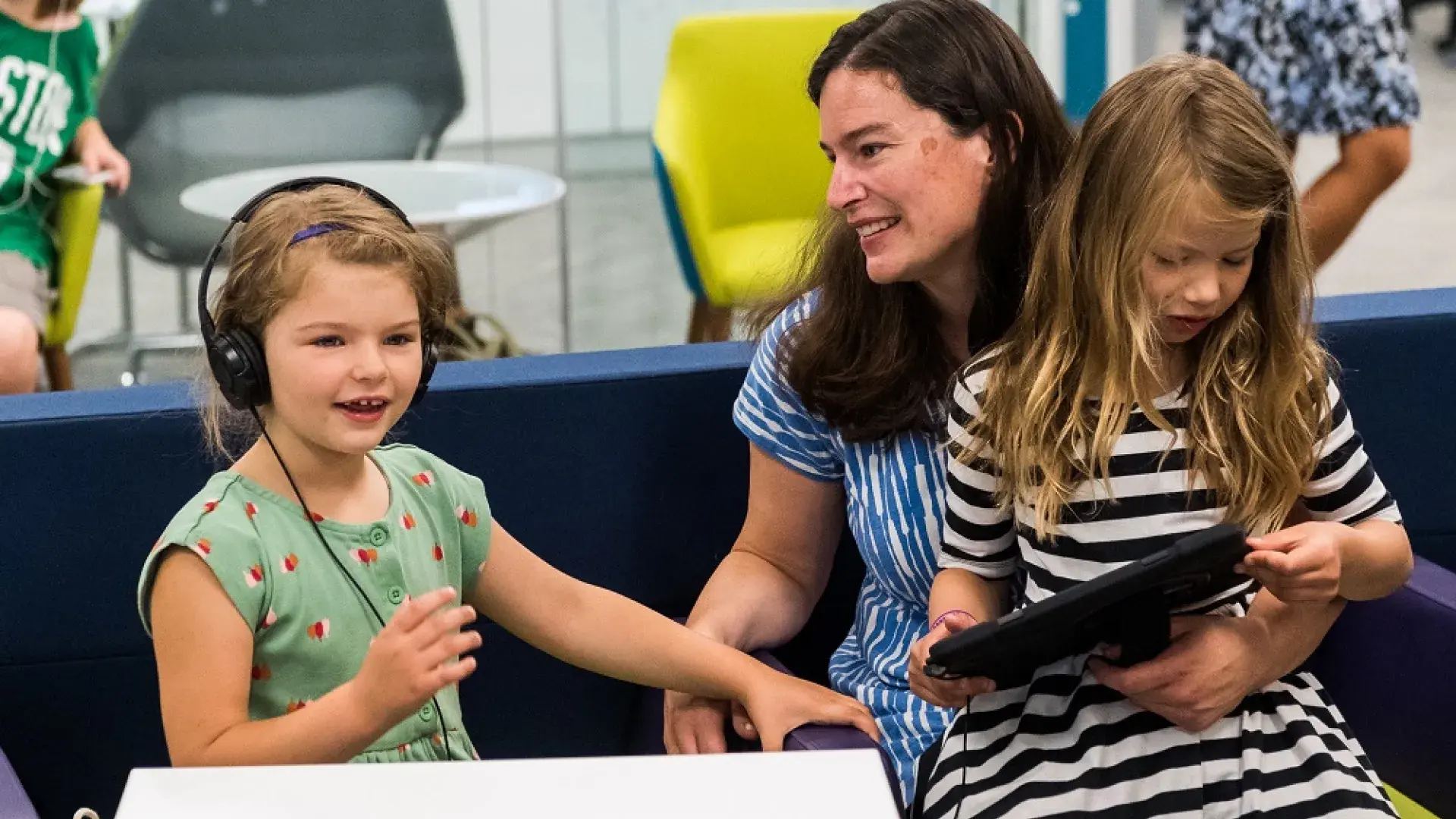 a little girl with headphones on sits next to a woman who has another little girl reading in her lap