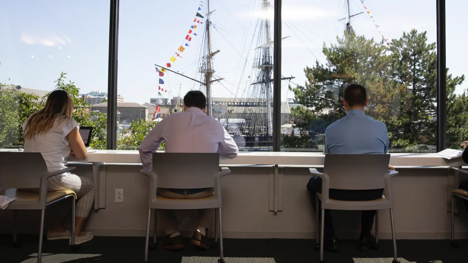 People working in front of a window with a colonial ship in the harbor