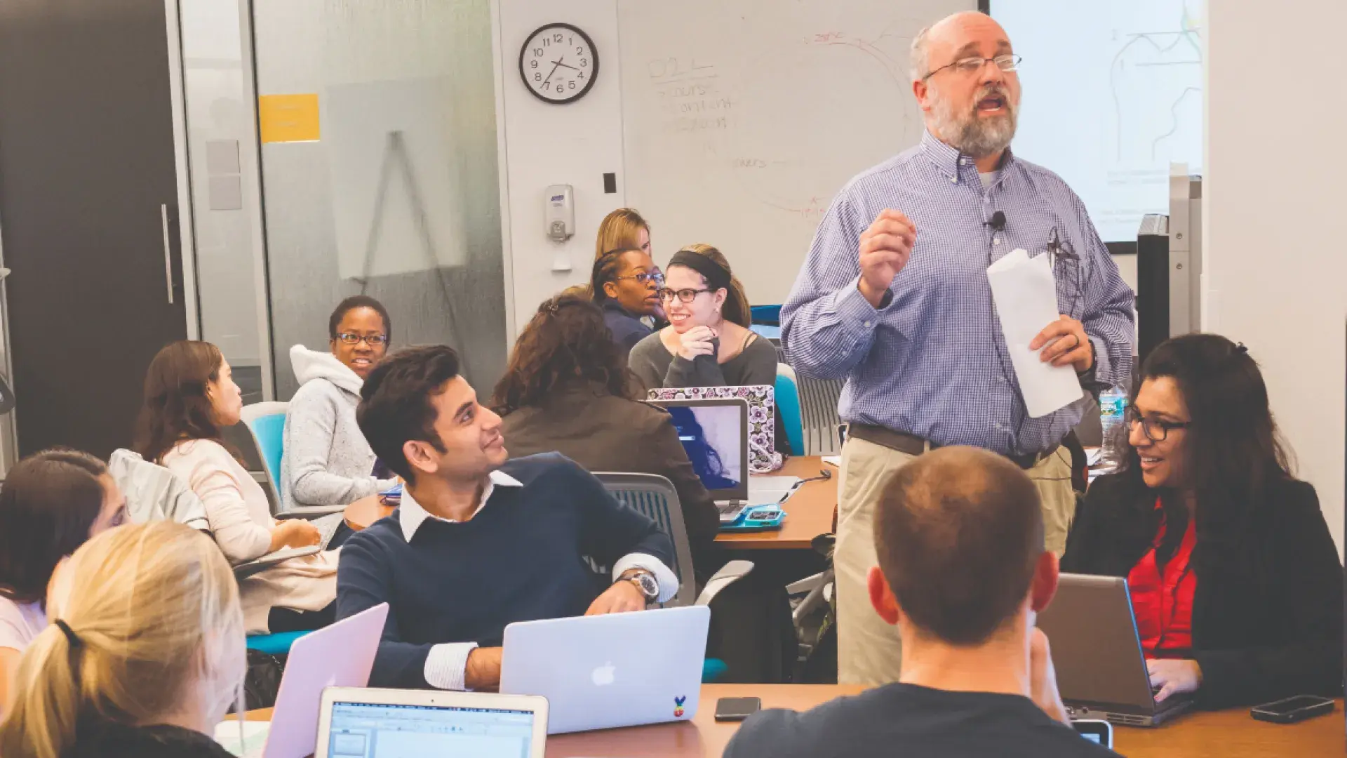 Male speaker addresses people on laptops seated at long tables to either side of him.