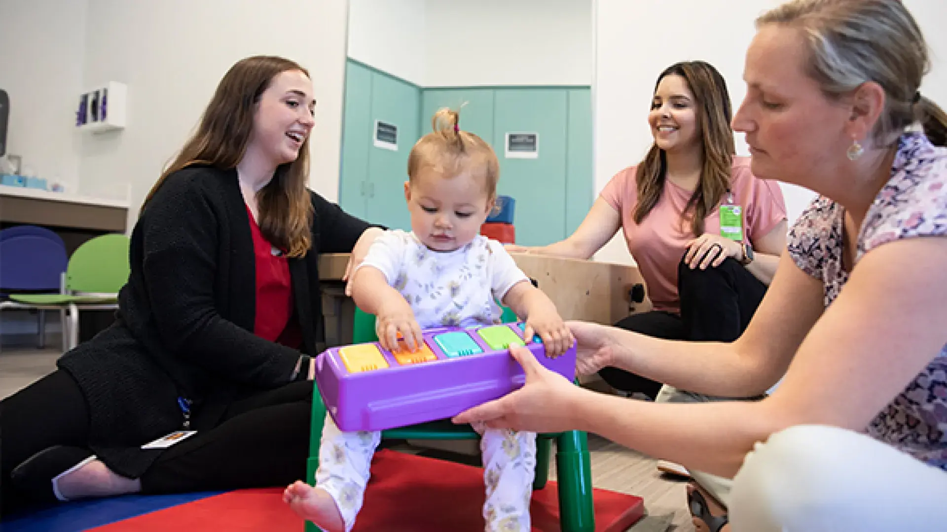 Three student women sitting on the ground smile and help a baby sitting on a small chair, handing it a colorful toy