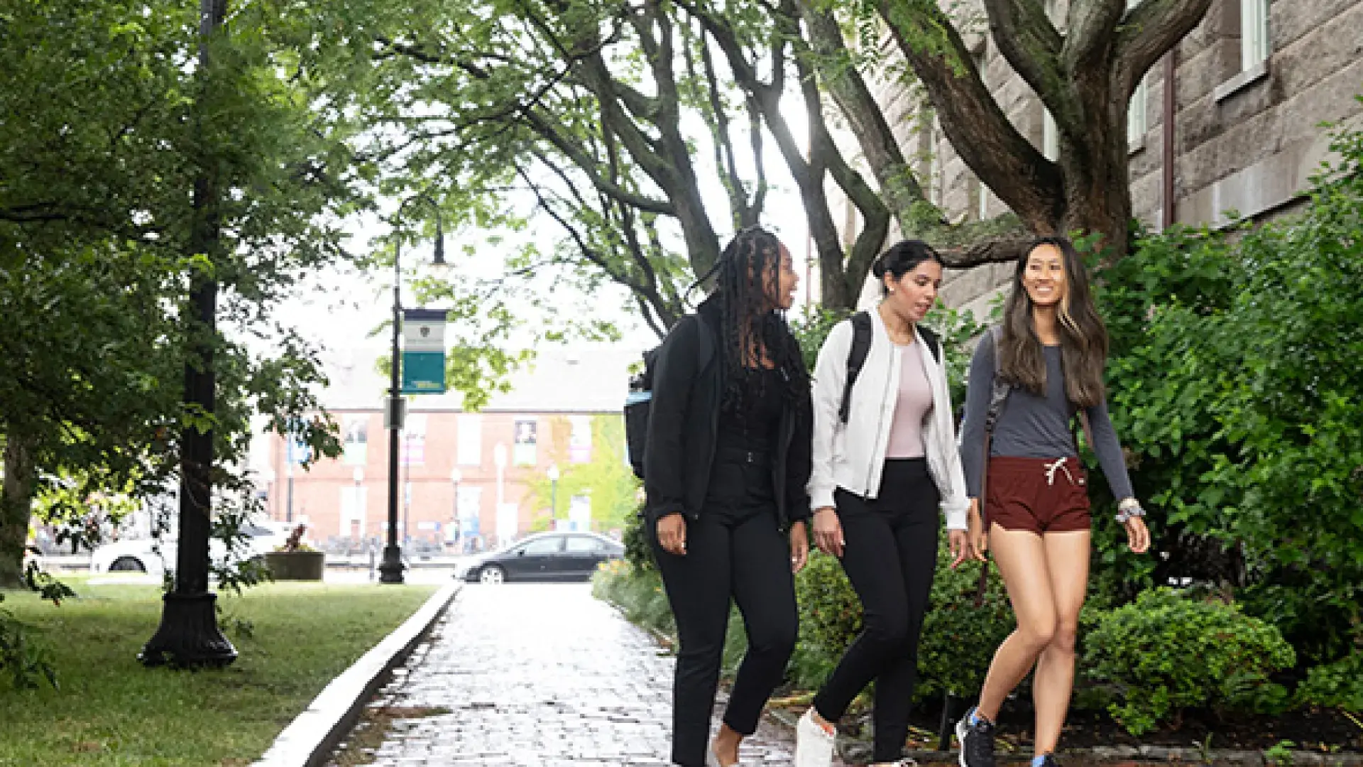 Three women walk on a stone path past trees and grass and lamp posts
