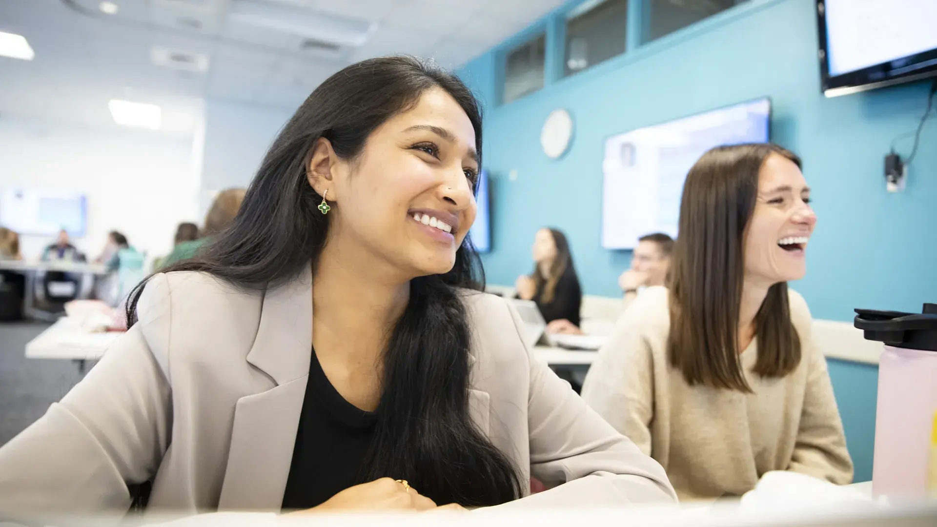 two females sit at a desk and laugh at something off camera