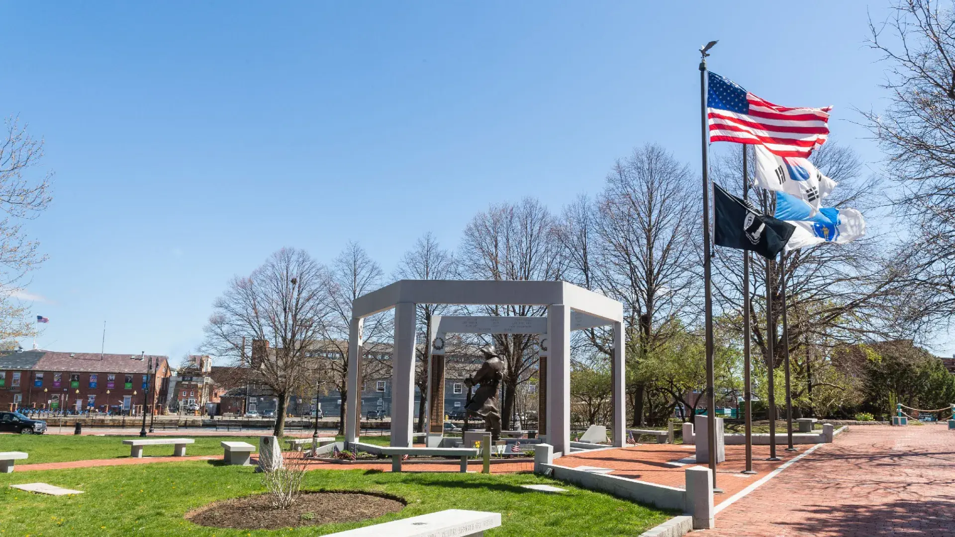 American Flag waves with POW MIA and other flags in front of a war memorial statue and a blue sky
