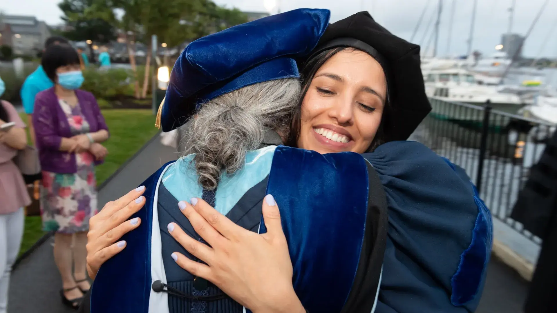 two people wearing dark blue graduation robes and hats hug happily 