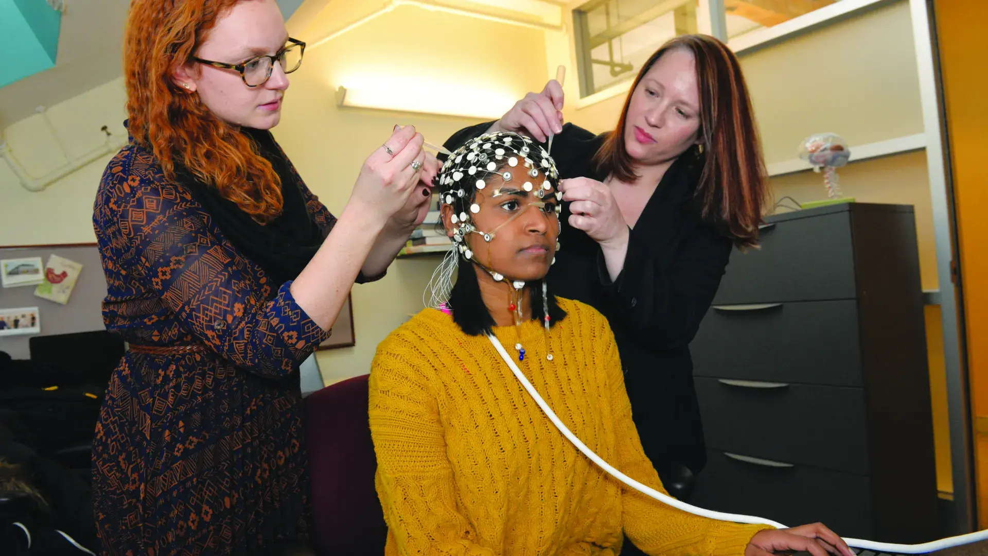 Two researchers place sensors on the head of a sitting person who is sitting and using a computer