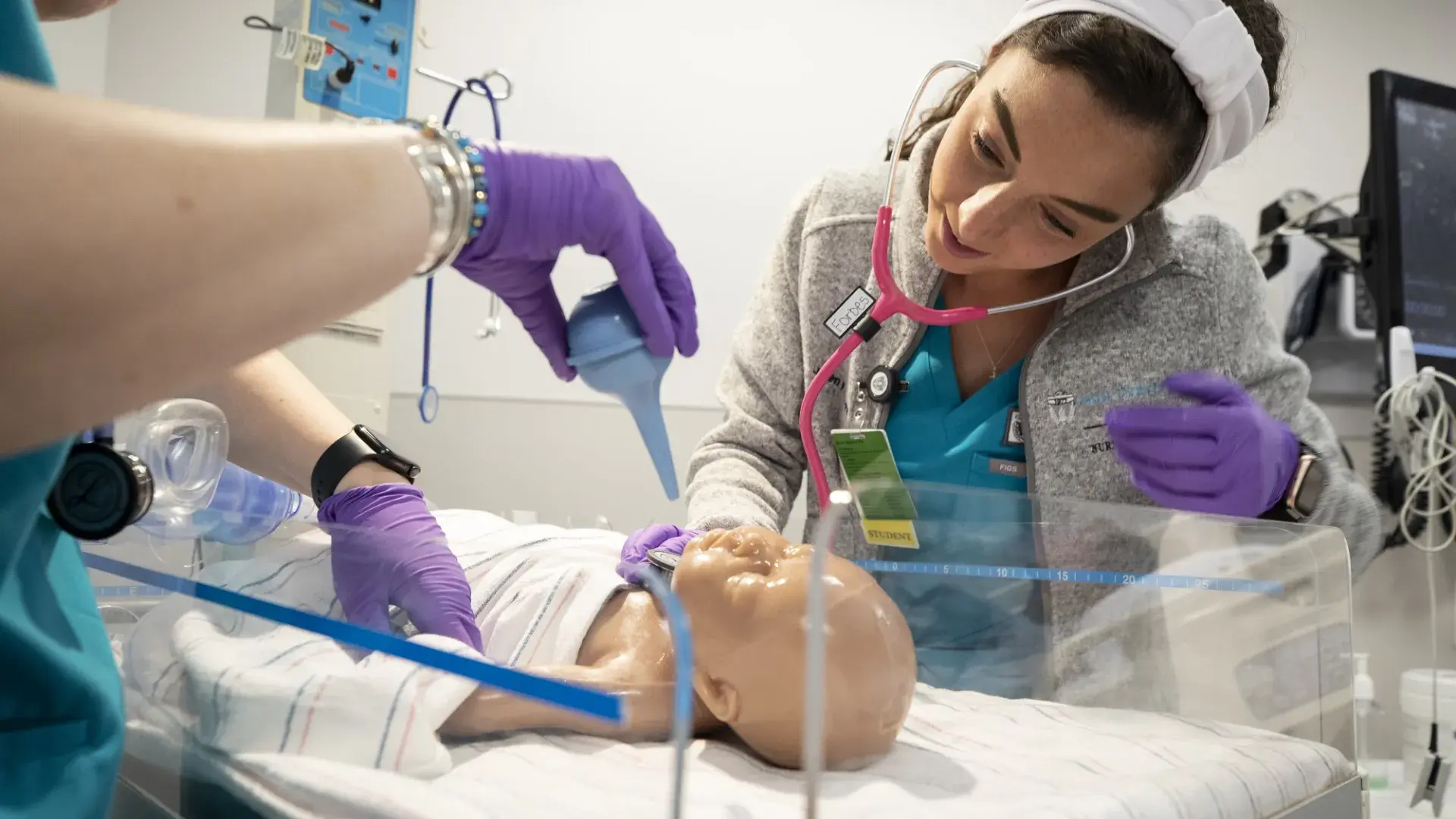 two women in scrubs look at a baby doll and use a suction bulb on it