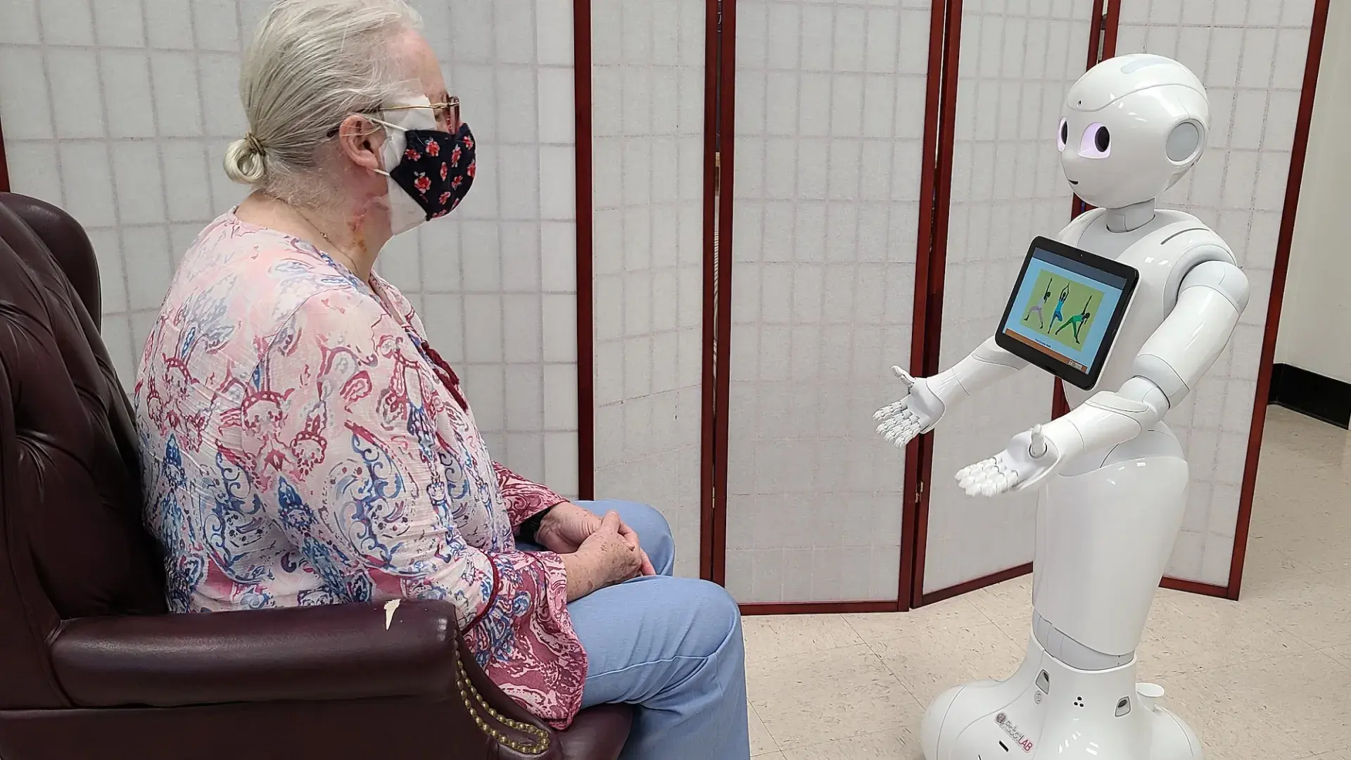a white android about 4 feet tall interacts with a seated elderly woman